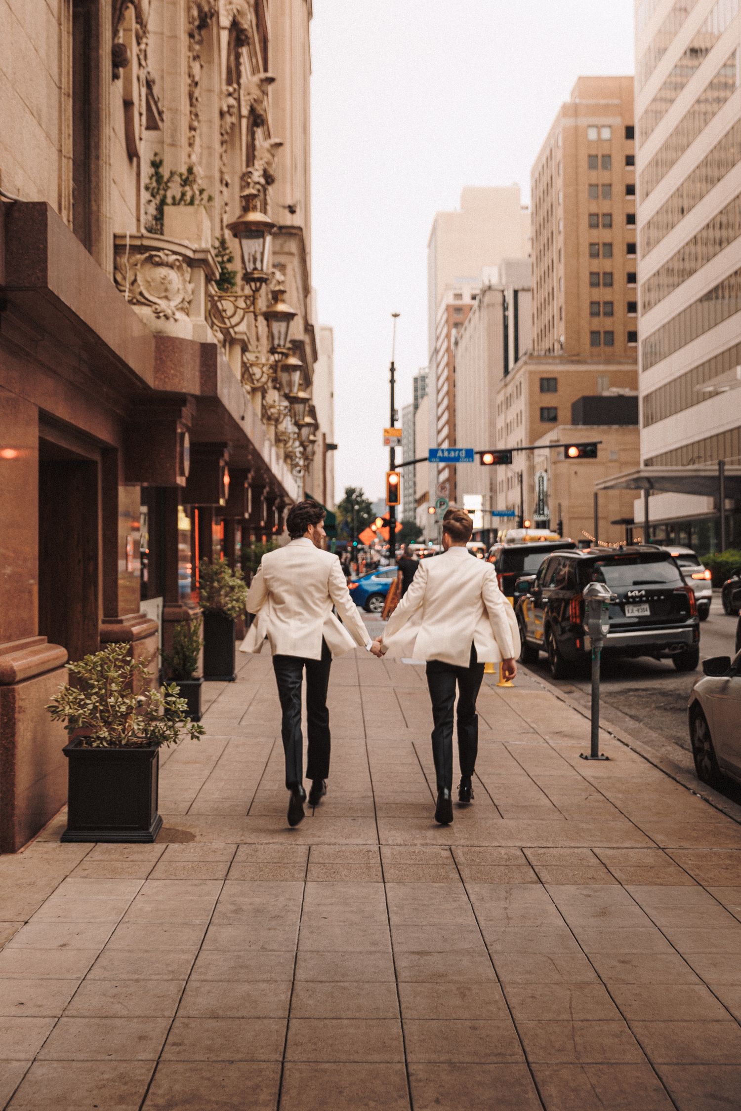 Two grooms in matching white tuxedos walking hand in hand down a city street.