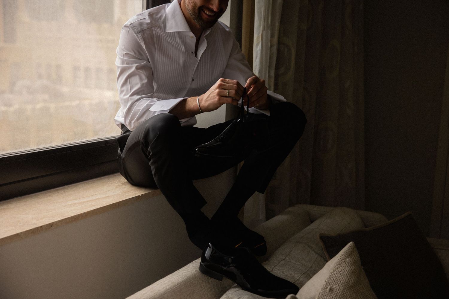 Groom adjusting cufflinks while seated on window ledge in white dress shirt.