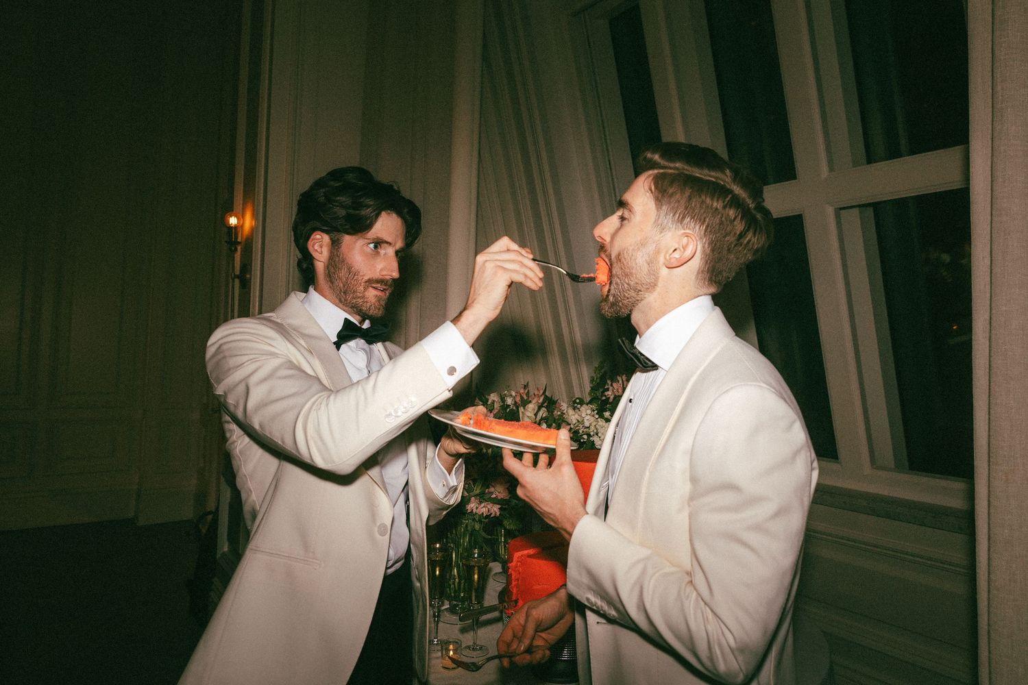 Two grooms in white tuxedos share a joyful cake-feeding moment at their wedding reception.