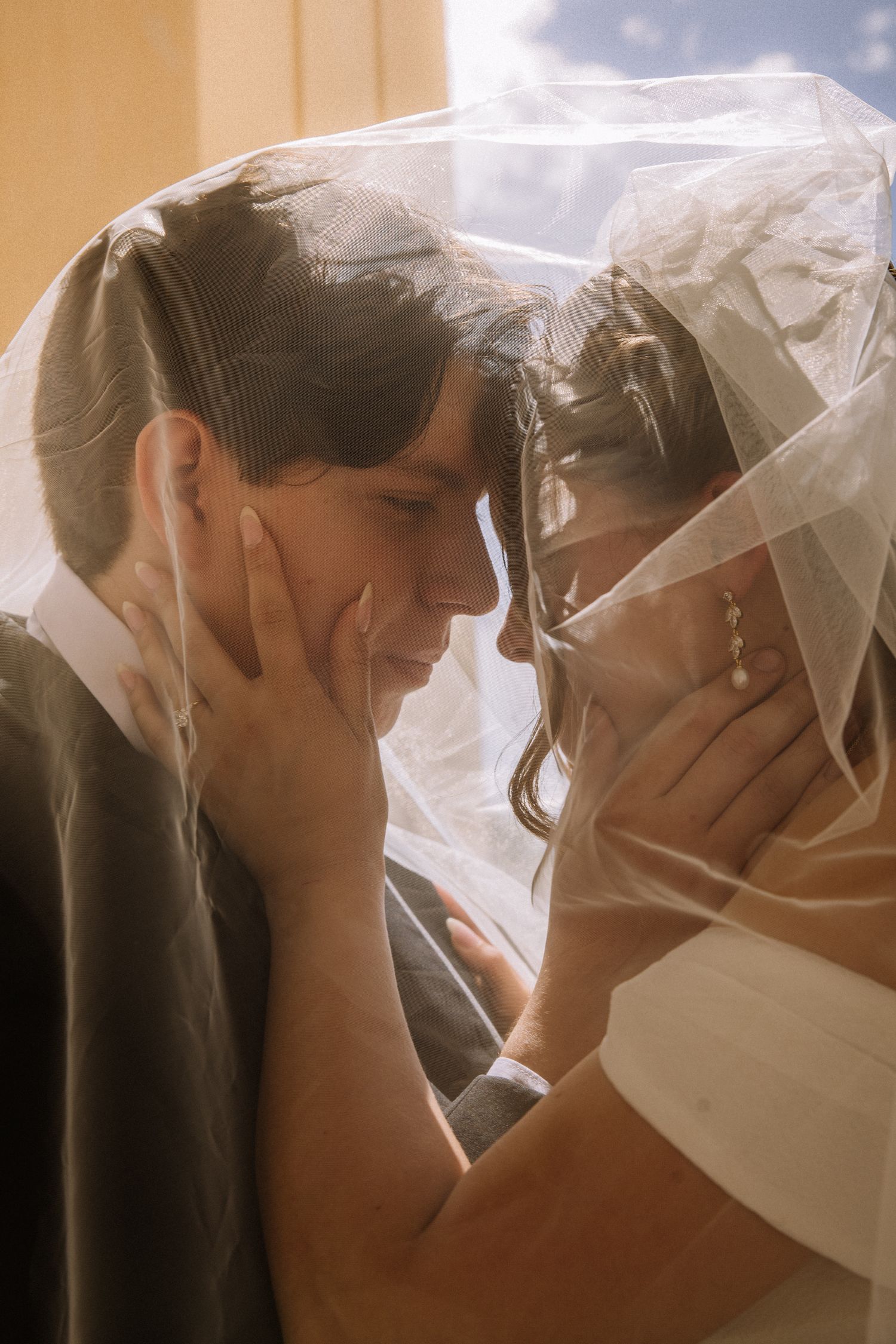 Bride and groom sharing intimate moment under flowing veil in soft golden light.