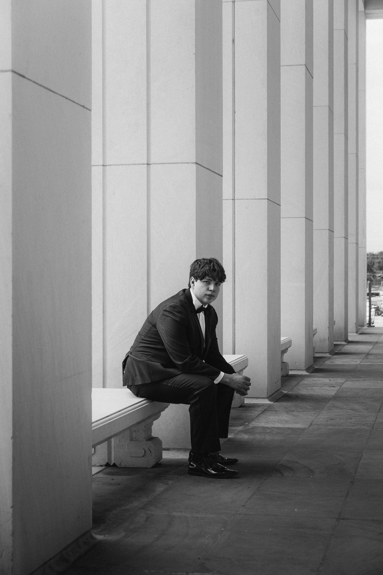 Groom in black tuxedo sitting pensively on stone bench between elegant columns in black and white photo.