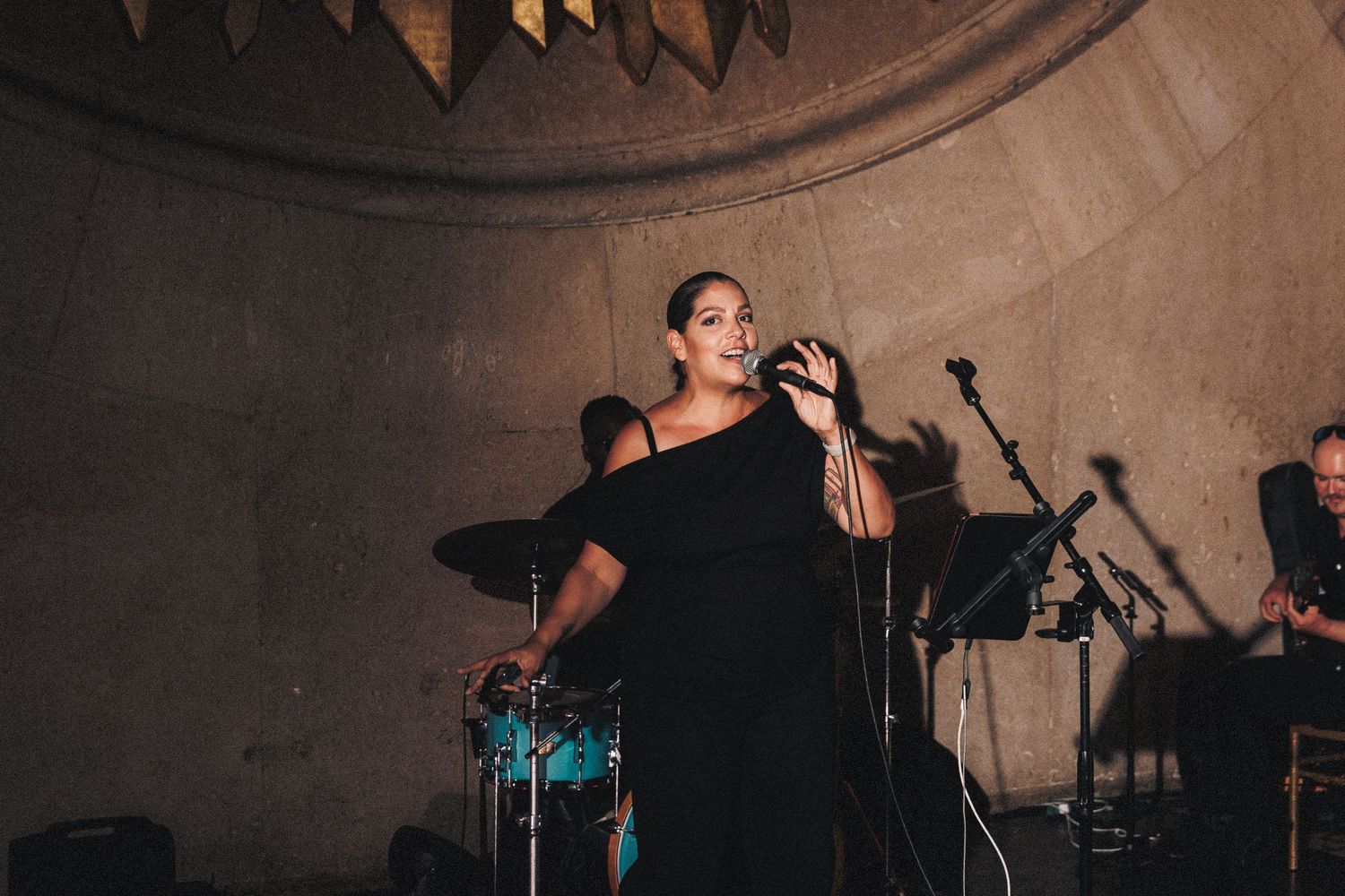 Woman in black off-shoulder dress singing on stage with band during live performance.