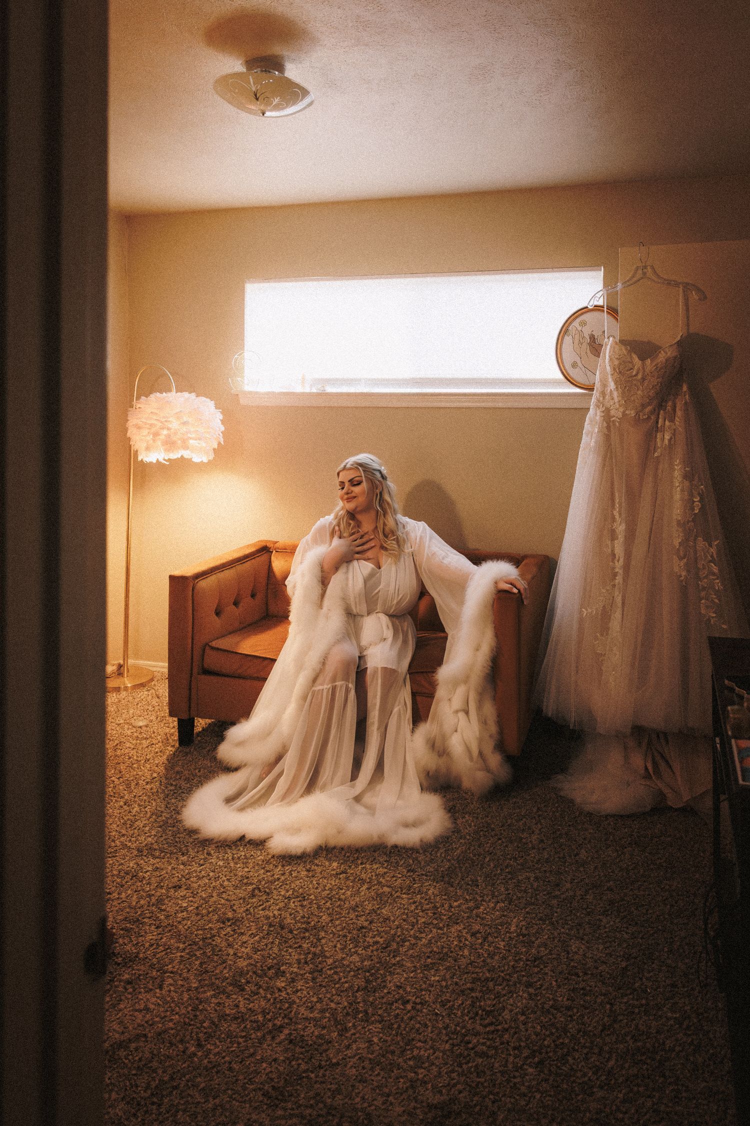 Bride in sheer fur-trimmed robe sitting near hanging wedding gown in warmly lit vintage bedroom.
