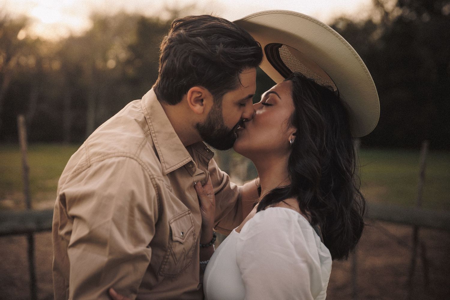 A couple sharing a romantic kiss outdoors at sunset, woman wearing a white cowboy hat and dress.