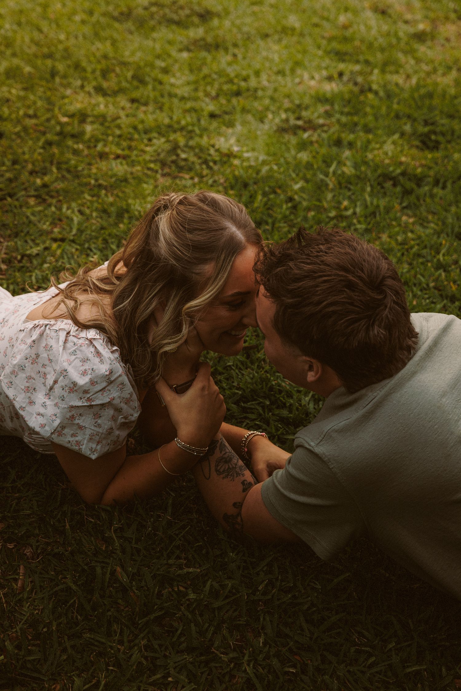 Couple lying on grass, faces close together, sharing a tender romantic moment outdoors.