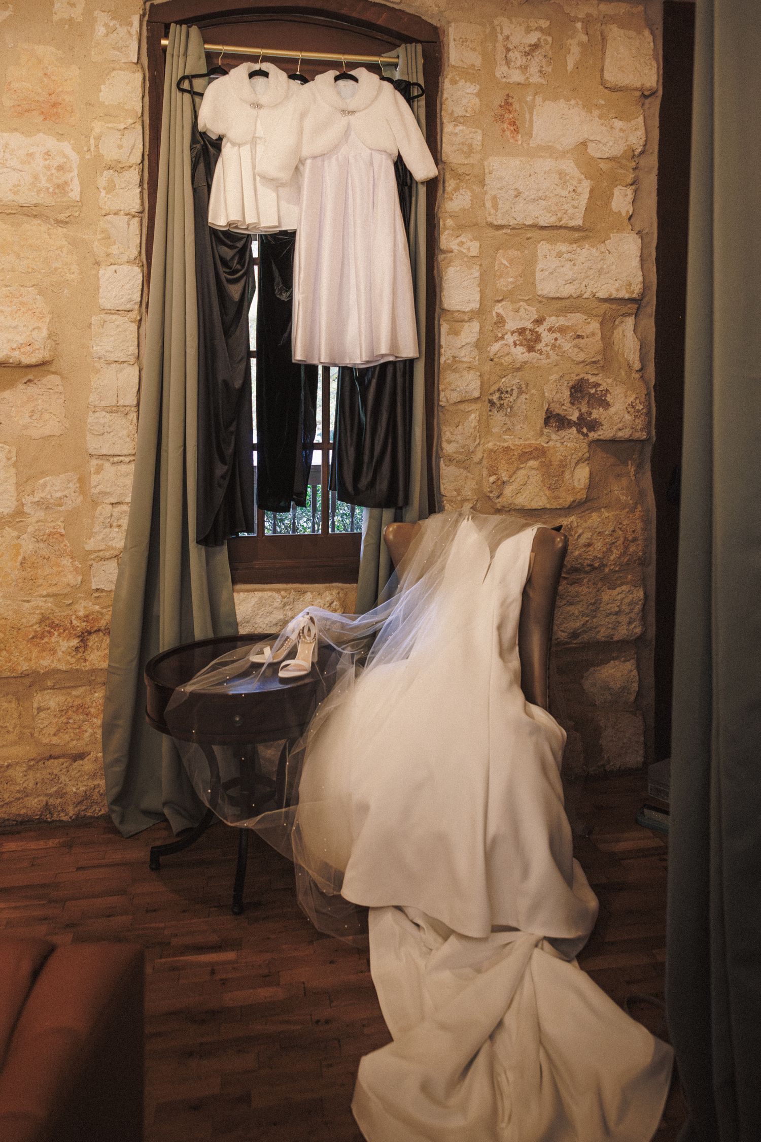 White wedding dress and flower girl dresses hanging in a rustic stone-walled bridal suite before the ceremony.