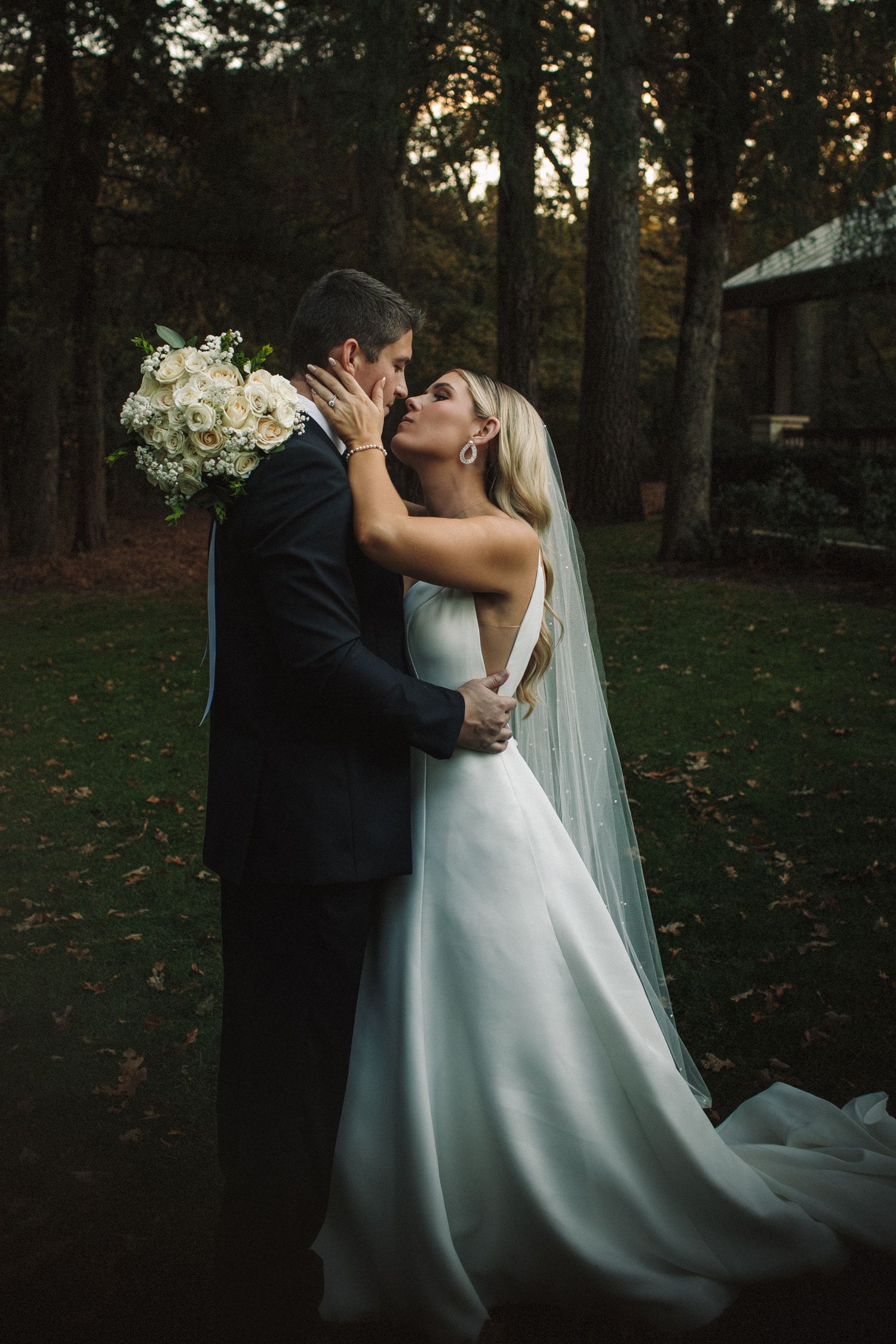 A bride and groom embrace and kiss while holding a white bouquet in a wooded outdoor setting at dusk.