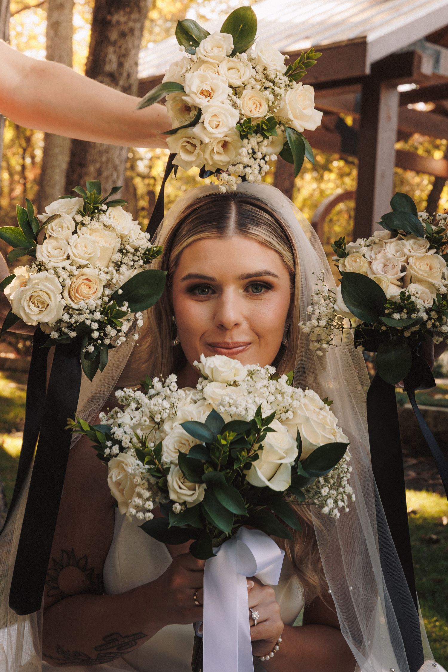 Bride surrounded by white rose bouquets with greenery and black ribbons, smiling on her wedding day.