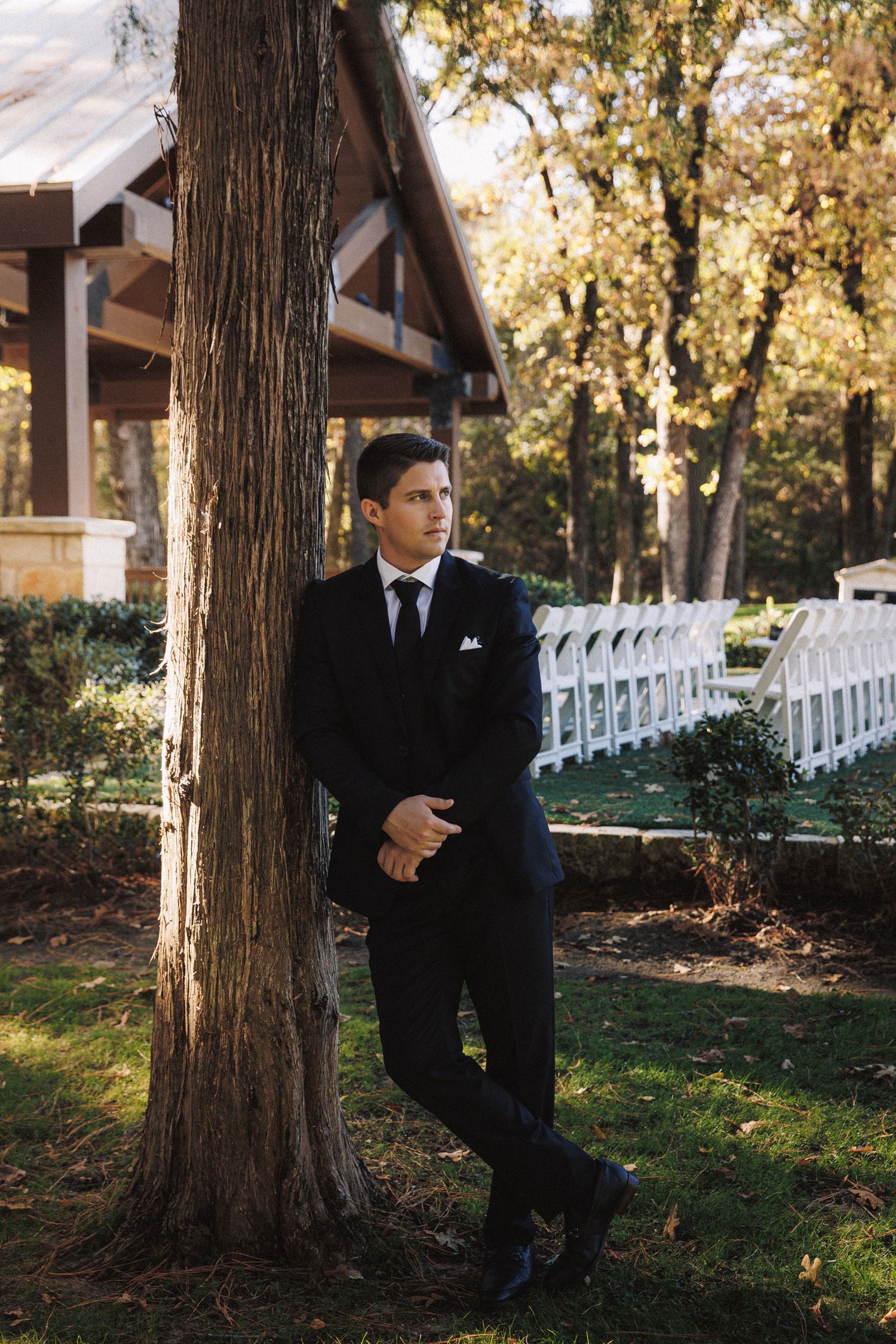 Groom in black tuxedo leaning against tree at outdoor wedding venue with white chairs.