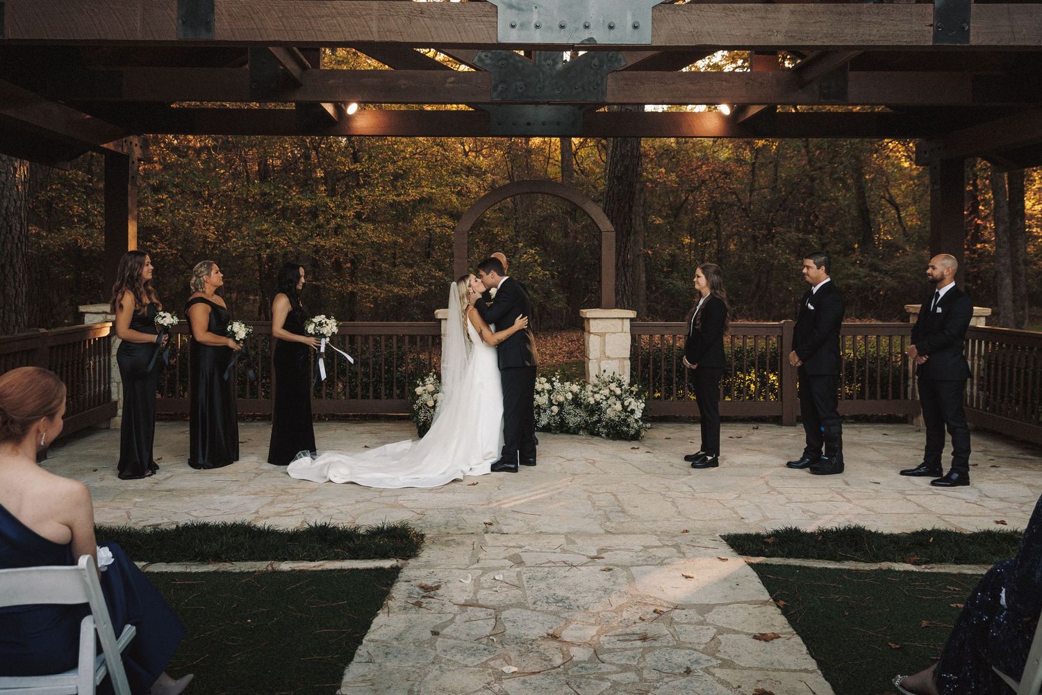 A bride and groom share their first kiss under a rustic pergola surrounded by bridesmaids and groomsmen at an outdoor wedding.