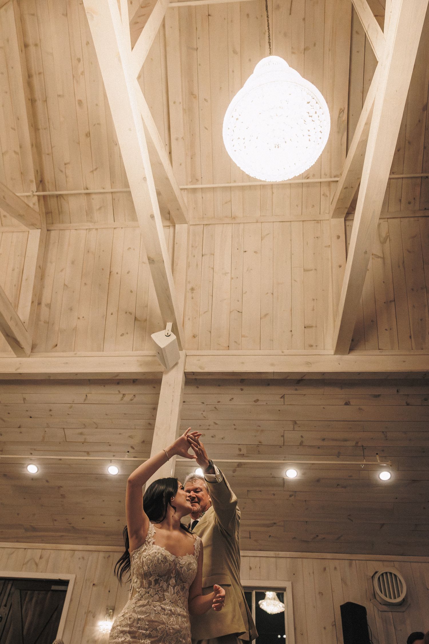 Bride in lace gown dancing with a partner under a decorative pendant light in a wood-beamed reception venue.