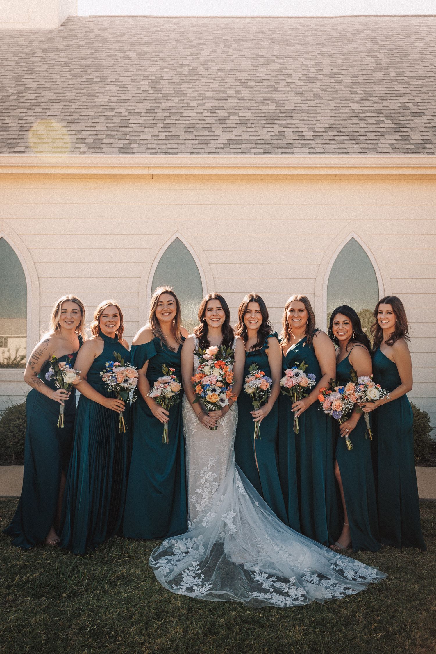 Bride in lace gown with seven bridesmaids in teal dresses holding bouquets outside a white chapel.