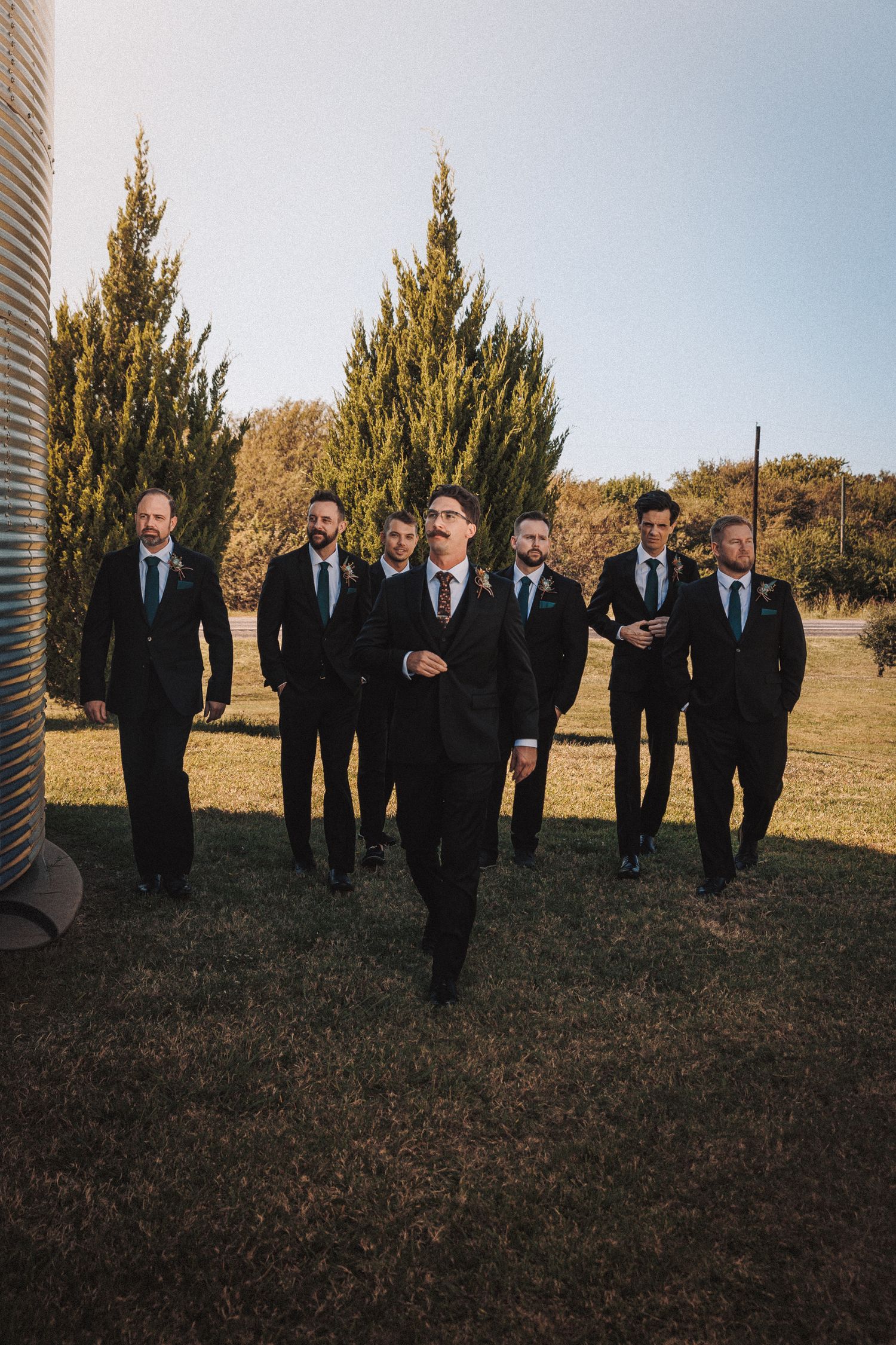 Groomsmen in black suits walking confidently outdoors near cypress trees on a sunny wedding day.