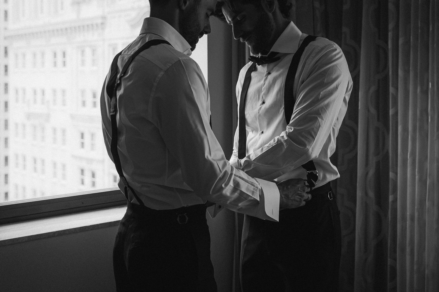 Black and white photo of two grooms in suspenders holding hands tenderly before their wedding ceremony.