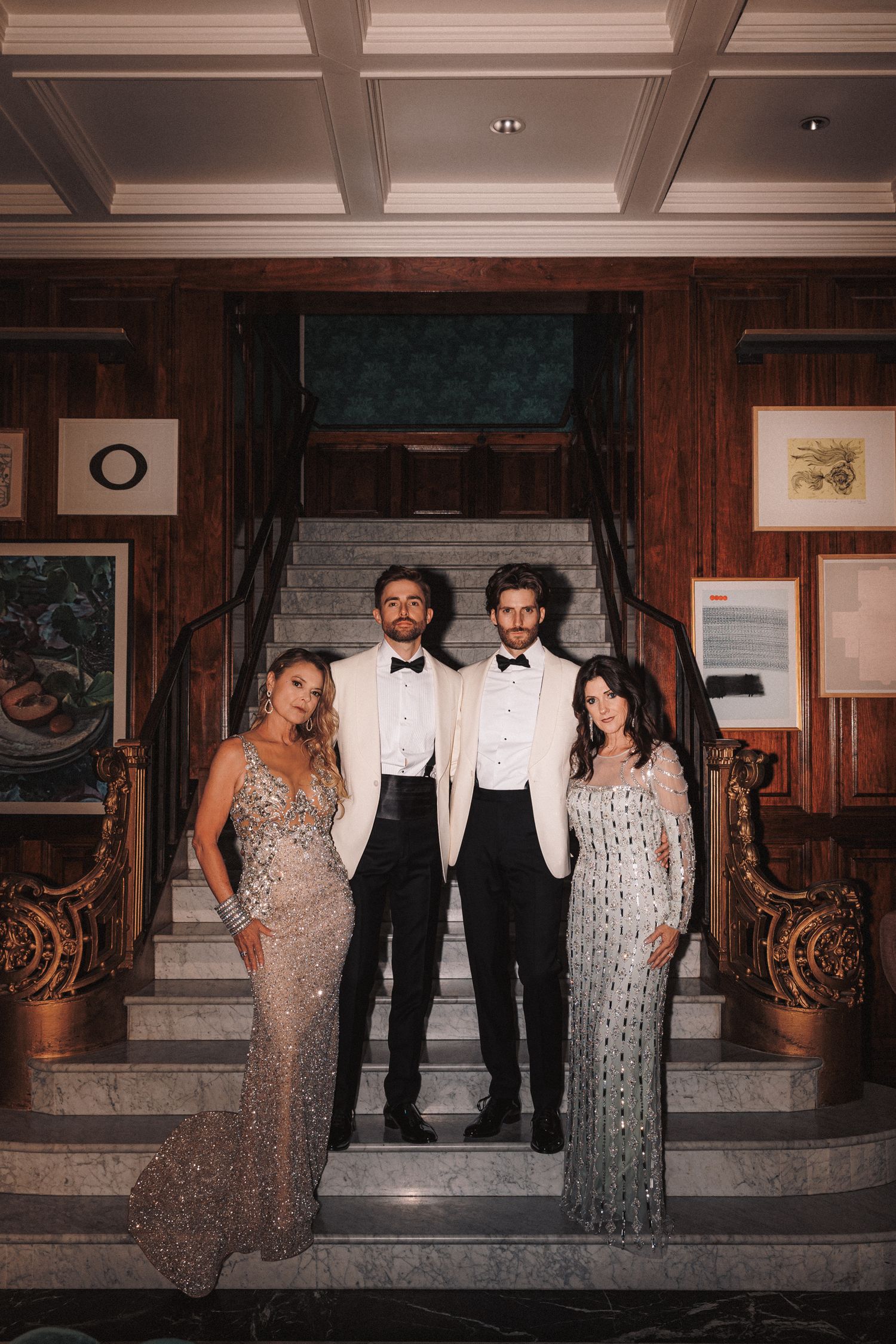 Two grooms in white tuxedos pose with two women in sequined gowns on elegant stairs.