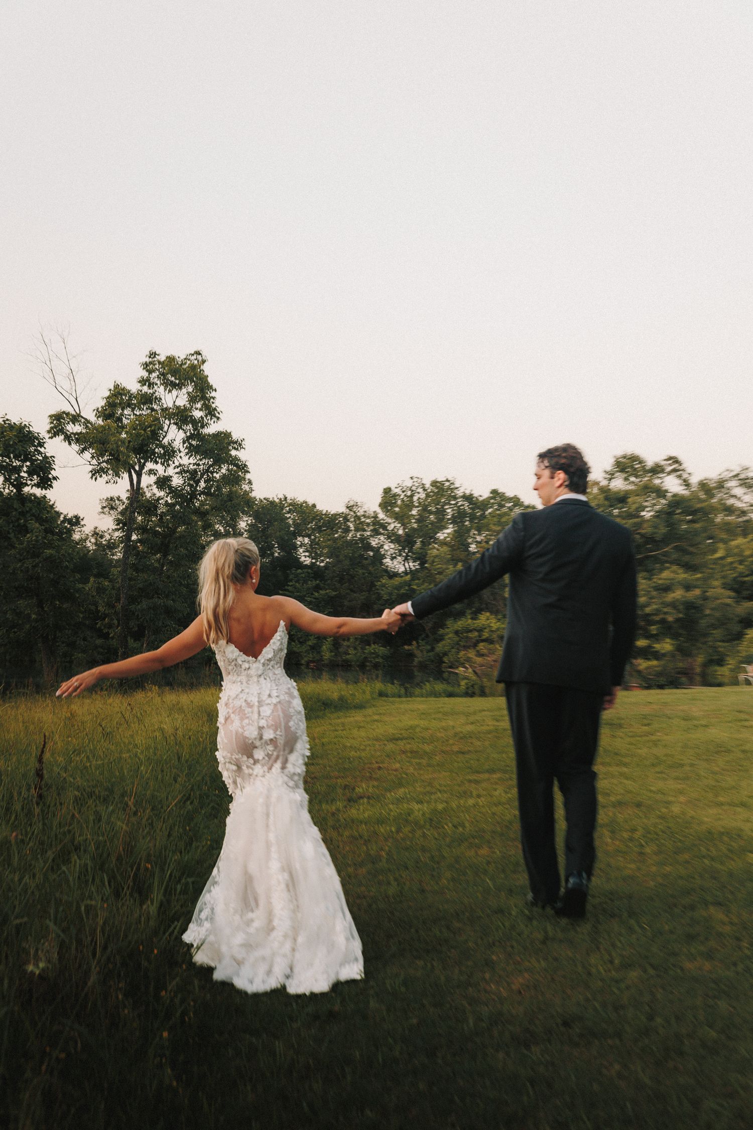 A newlywed couple poses in a grassy field during sunset, creating romantic silhouettes against the evening sky.