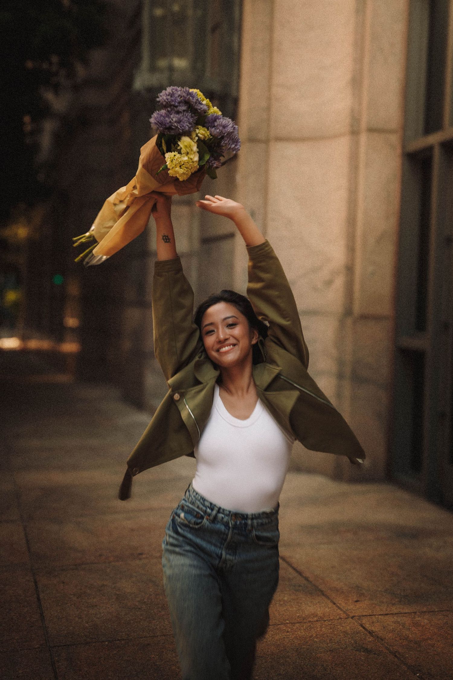 A woman in a white tank top and olive jacket twirls playfully in an urban setting with stone columns.