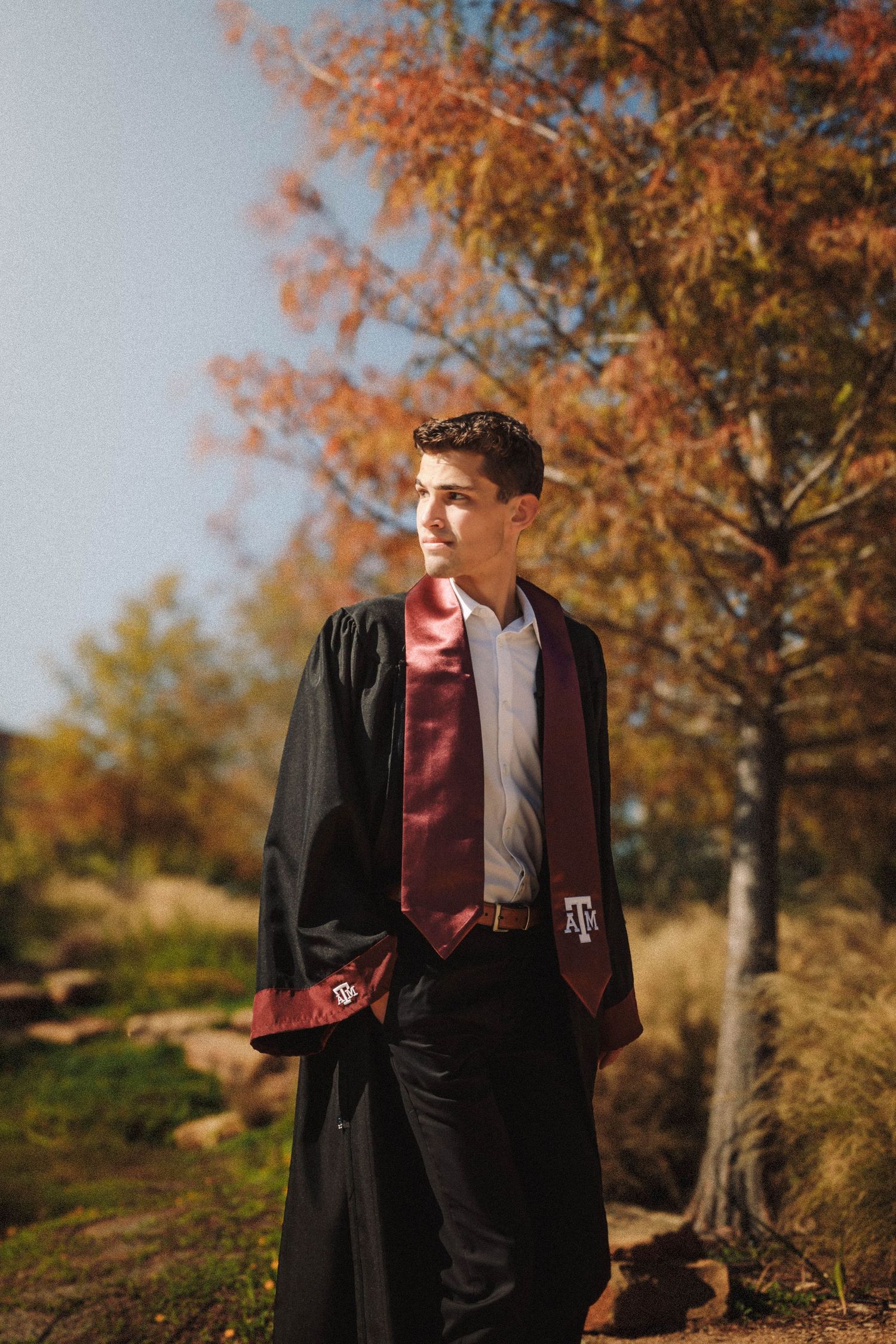 A graduate in a black gown and burgundy stole poses against autumn trees with golden leaves during a fall photoshoot.
