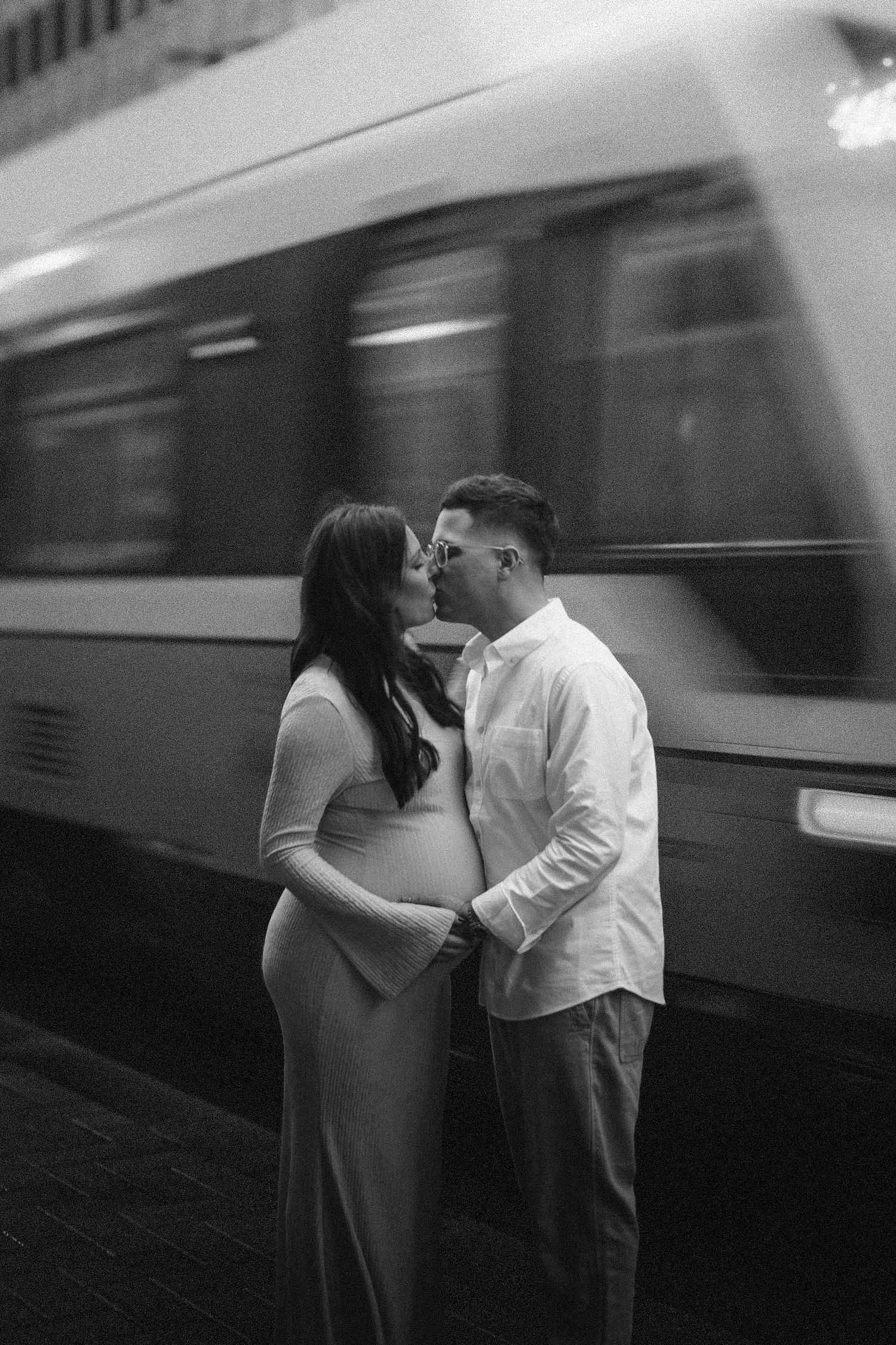 Black and white photo of expecting couple sharing intimate moment at train station as train passes in motion blur.