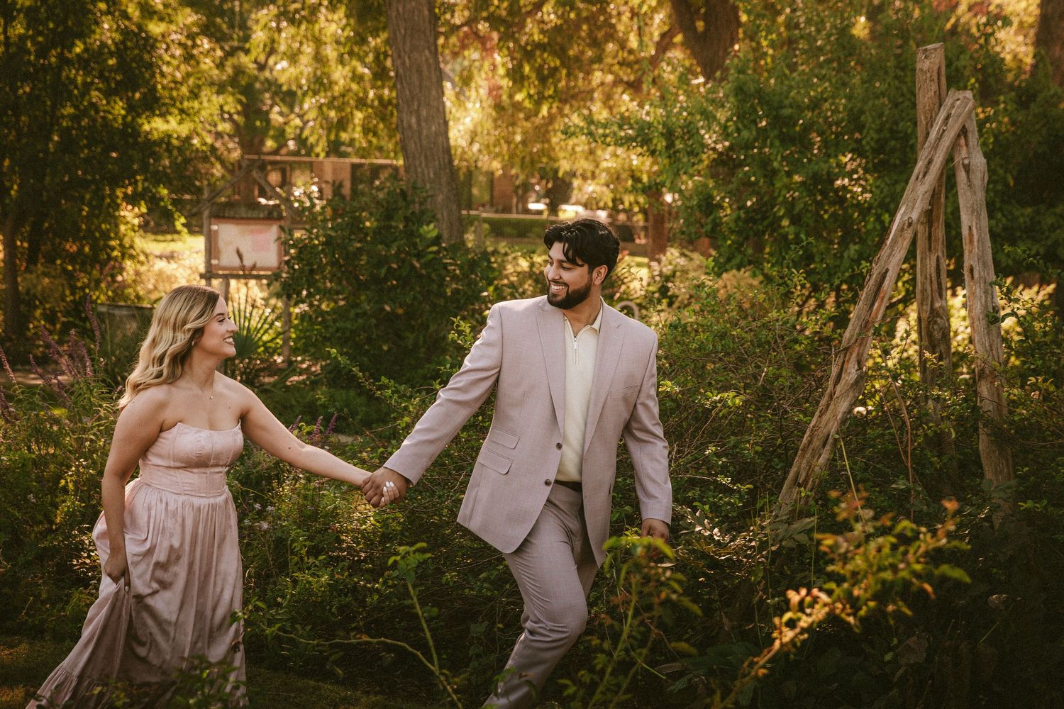 A couple in coordinated blush outfits walks hand in hand through a sunlit garden path.