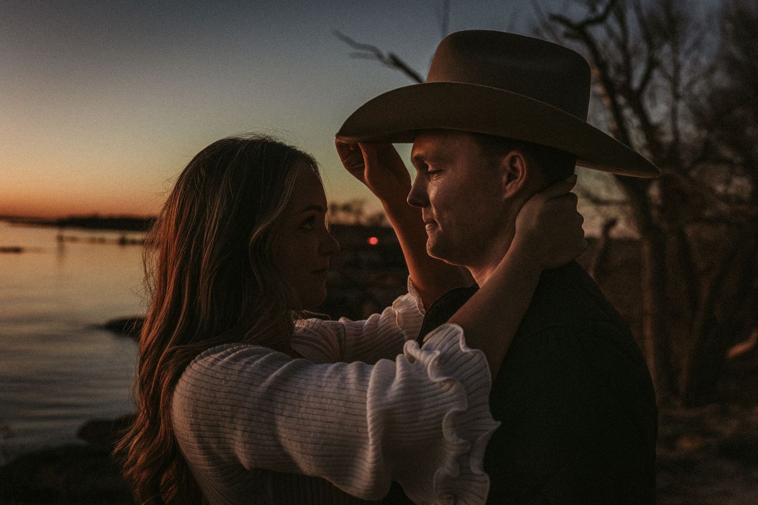 Silhouetted figures embrace during sunset with water and trees in the background.