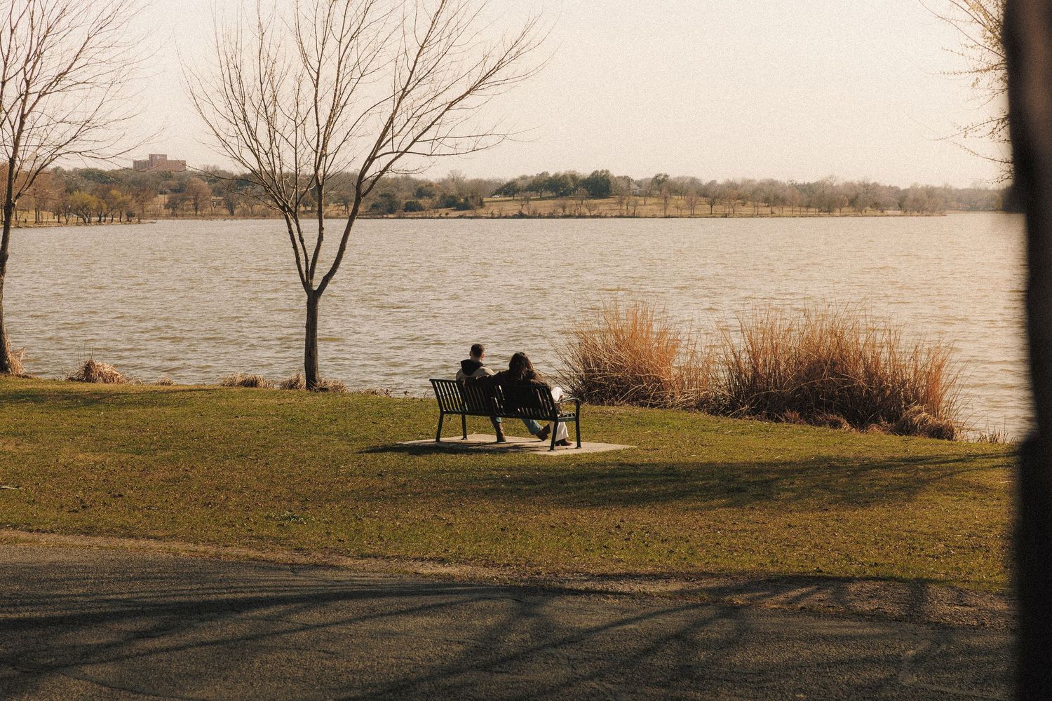 Silhouettes of two people sitting on a bench by a serene lake surrounded by bare trees.