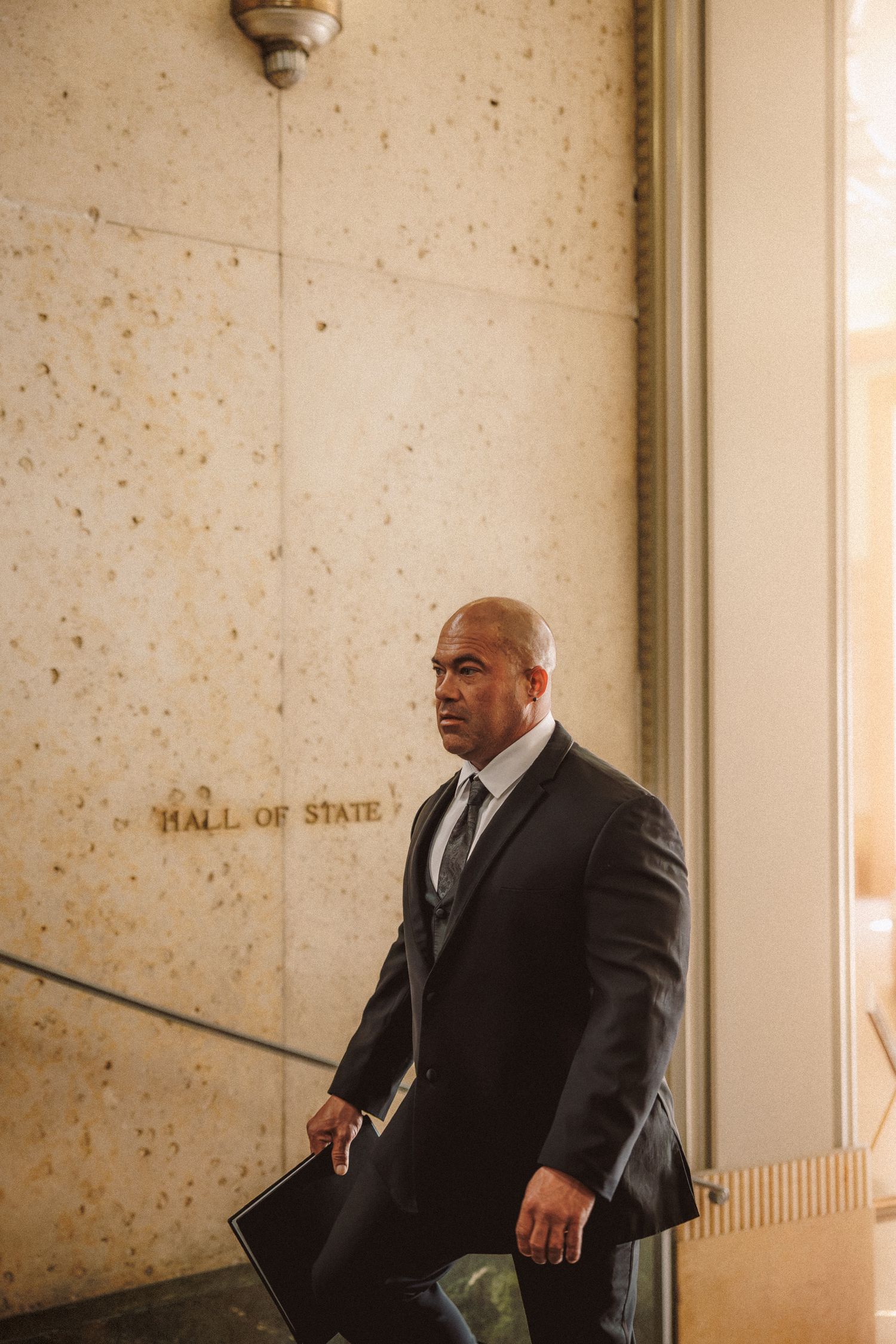 Bald man in dark suit carrying folder walks past Hall of State engraved marble wall.