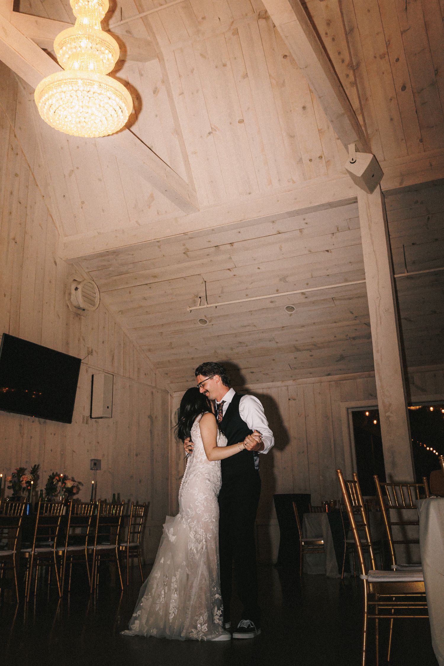Bride and groom share first dance under a chandelier in a rustic wood-paneled barn venue.