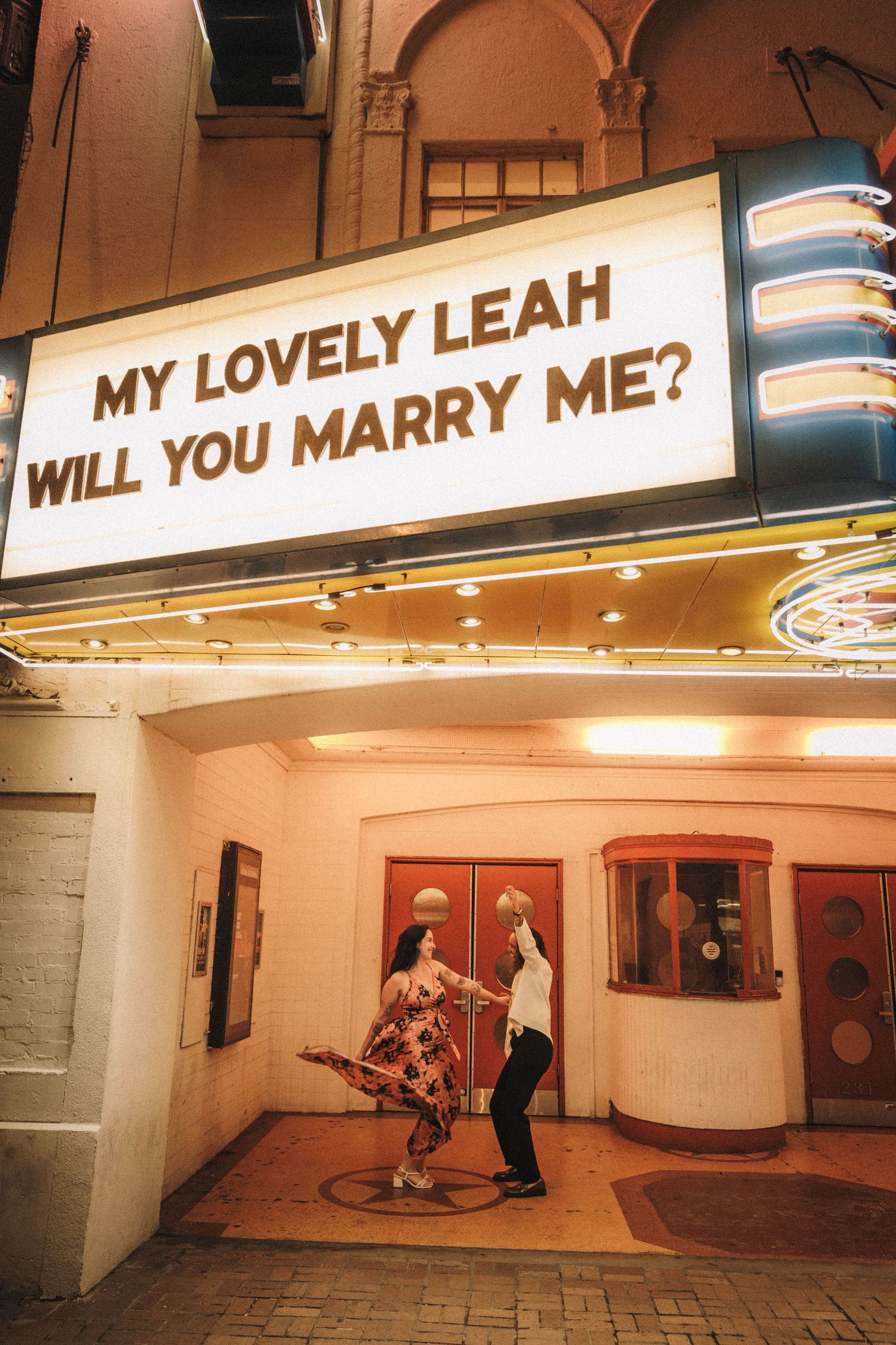 Couple dancing joyfully under a lit marquee reading My Lovely Leah Will You Marry Me.