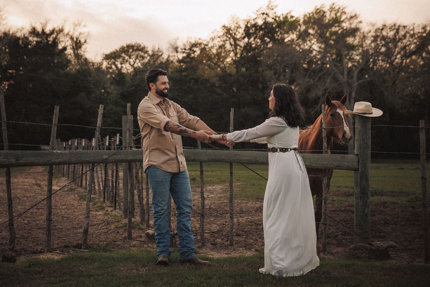 Engaged couple holding hands near a rustic wooden fence with a horse at golden hour on a farm.