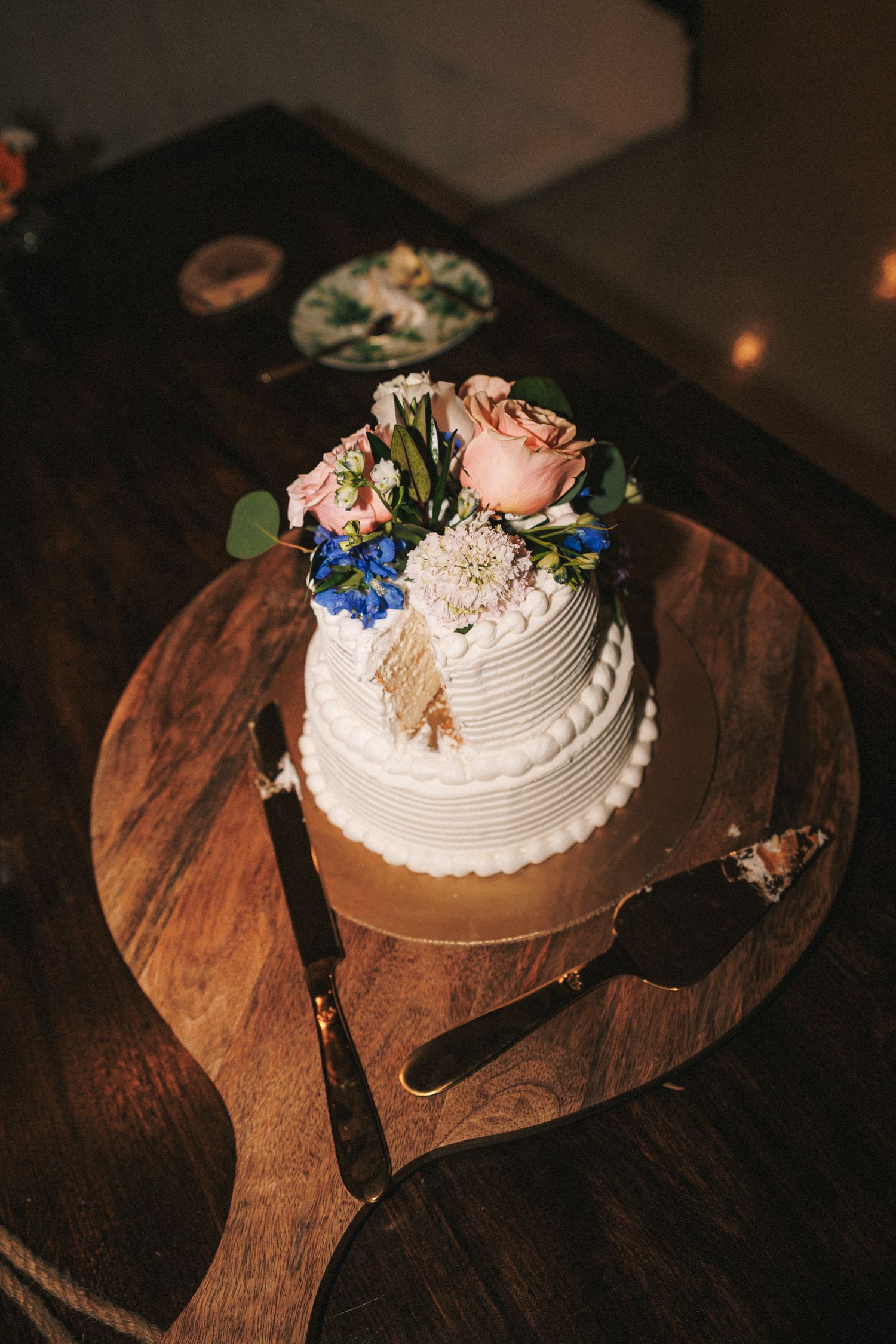 Elegant two-tier white wedding cake topped with pink roses and blue flowers on rustic wooden board.