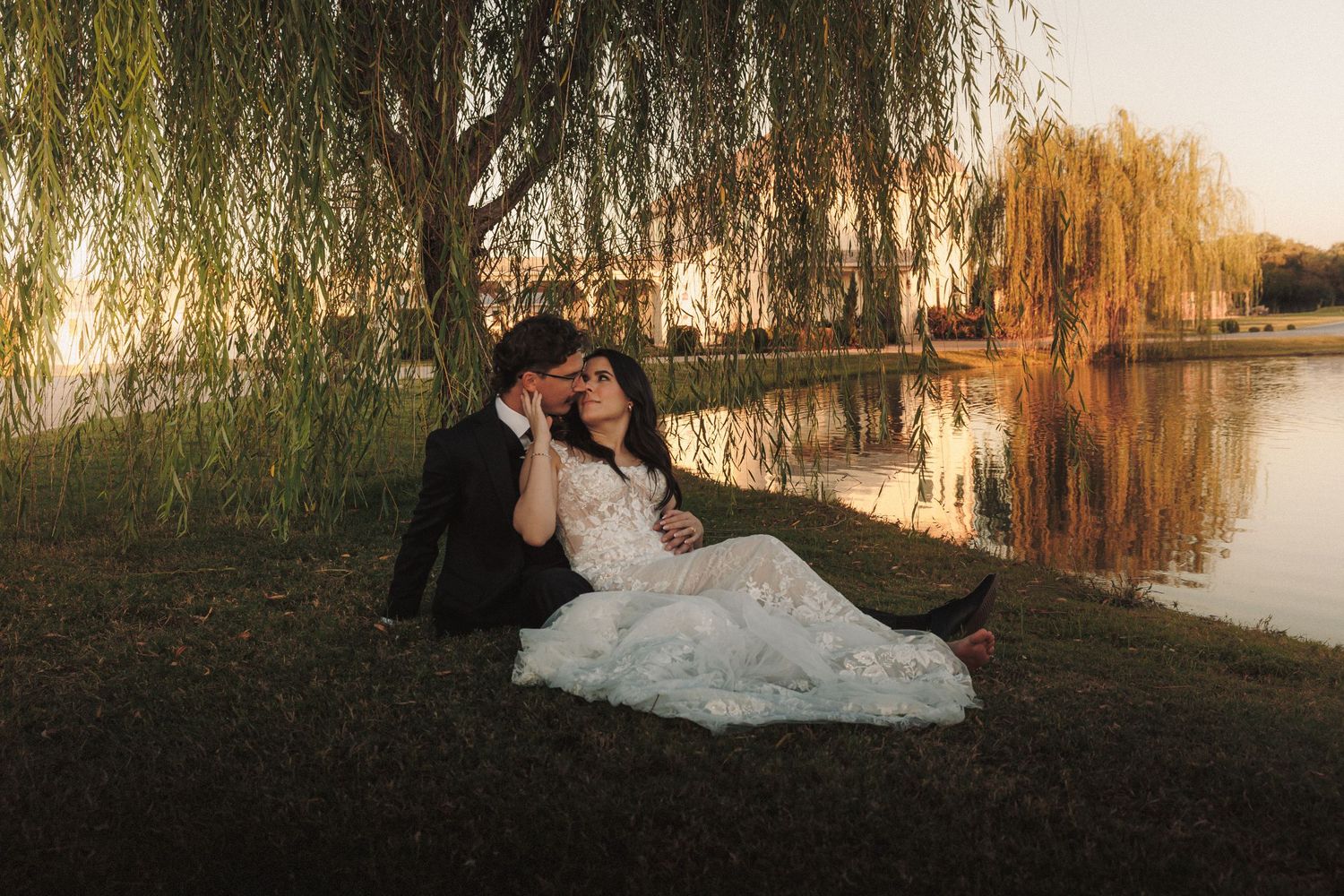 Bride and groom sitting together under a weeping willow tree by a reflective lake at sunset.