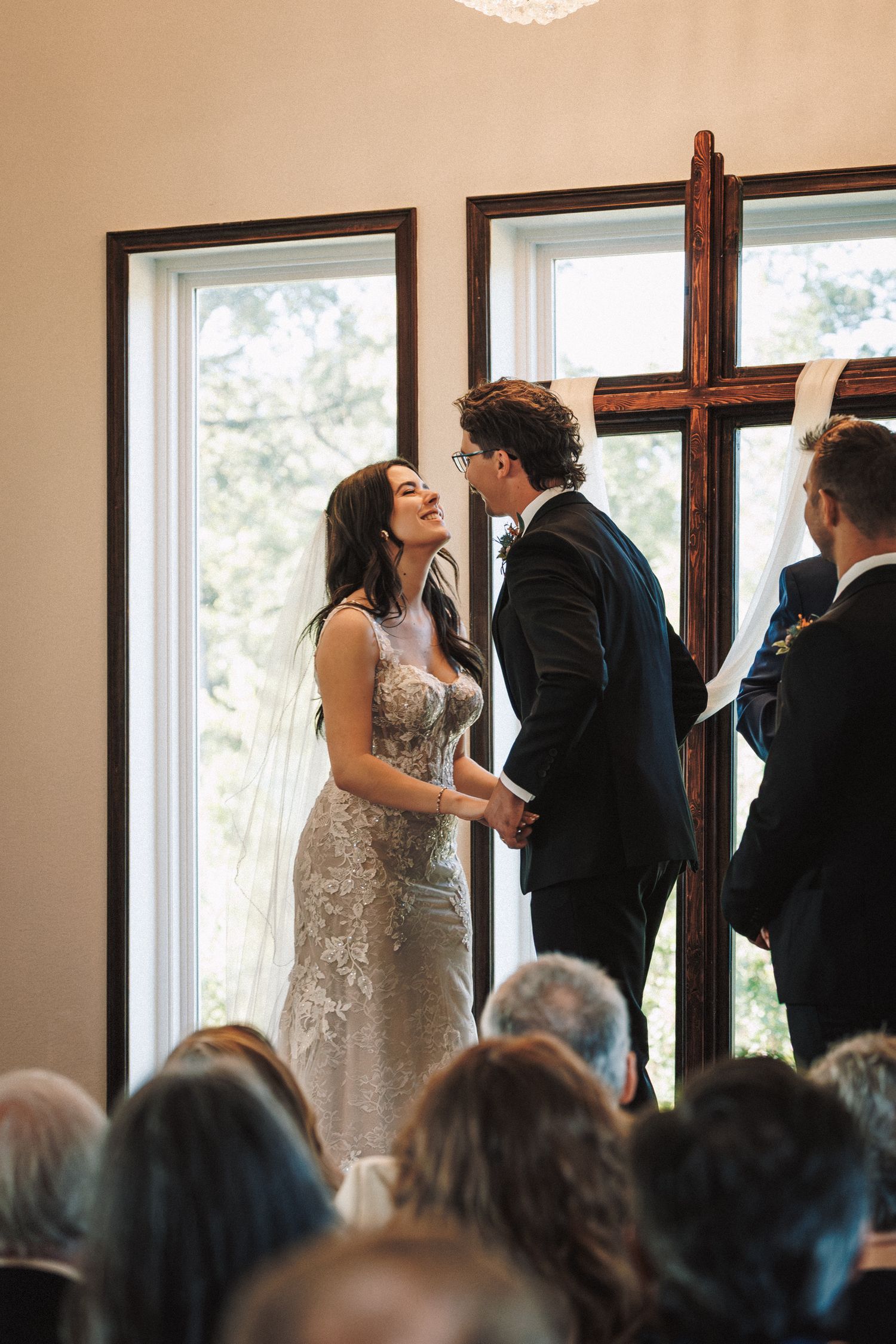 Bride and groom holding hands at indoor wedding ceremony before cross with guests seated below.