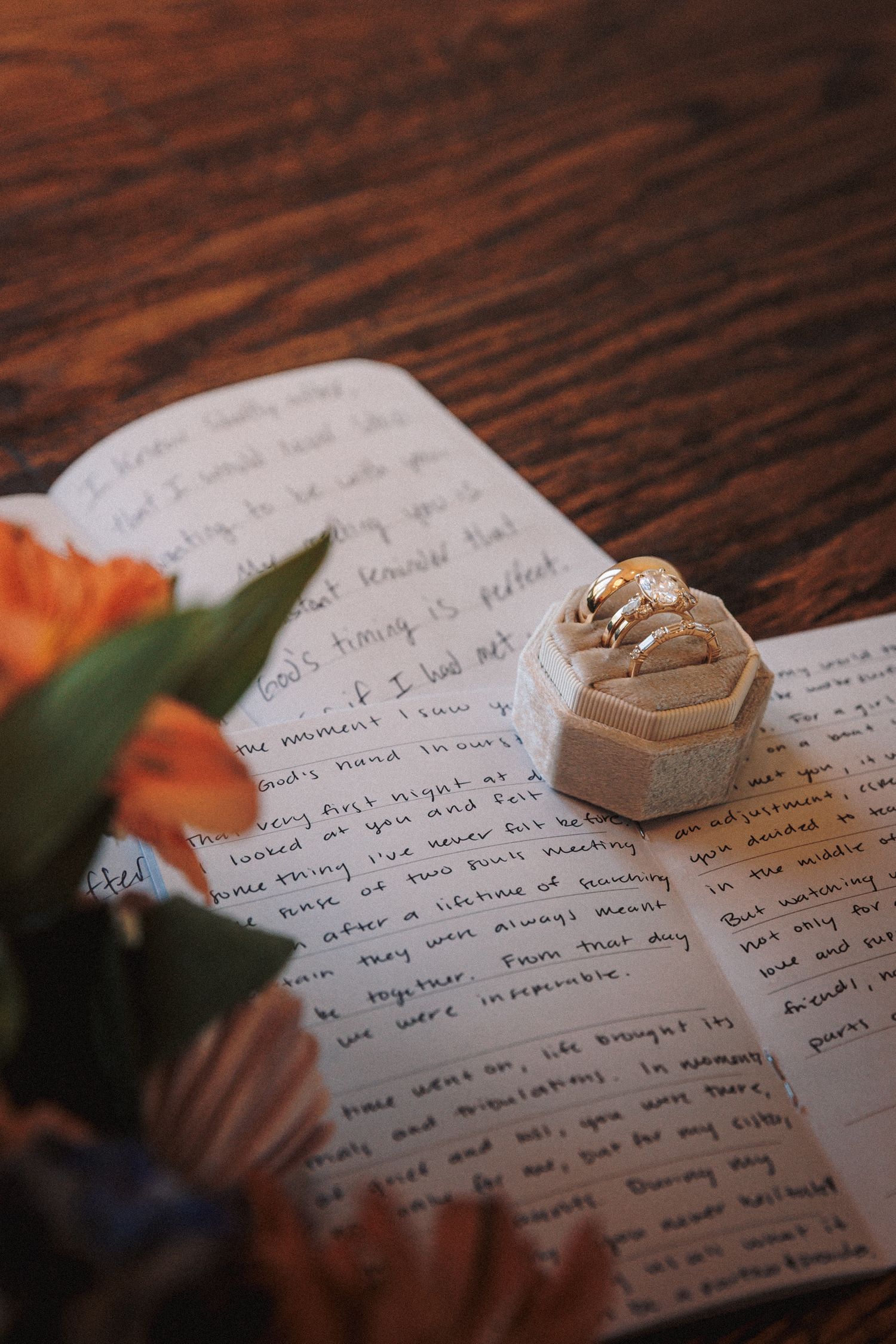 Wedding rings in velvet box resting on handwritten vows journal with orange flowers.