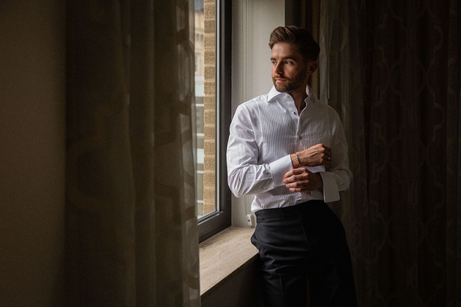 Groom in white dress shirt fastening cufflinks while standing by window in soft natural light.