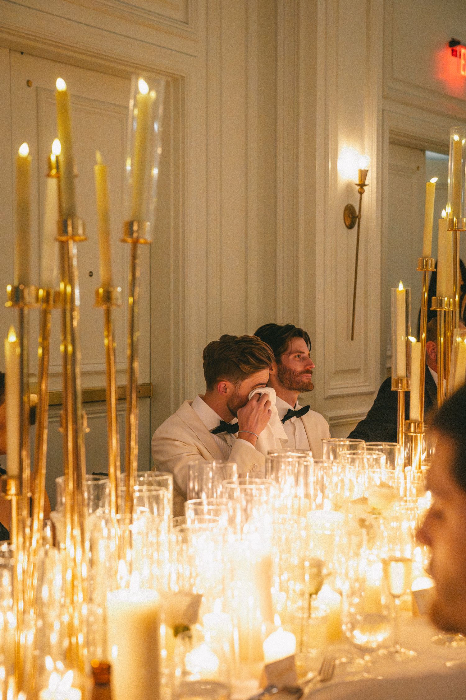 Two grooms sitting at a candlelit wedding reception table, one wiping tears during speeches.