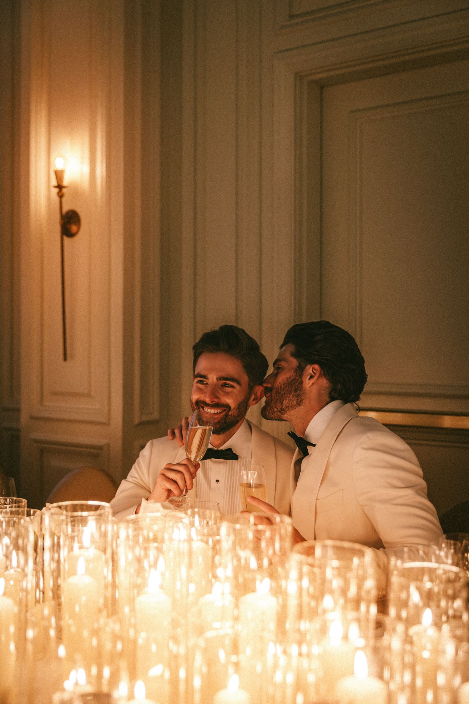 Two grooms in ivory tuxedos sharing a joyful candlelit moment at their wedding reception sweetheart table.