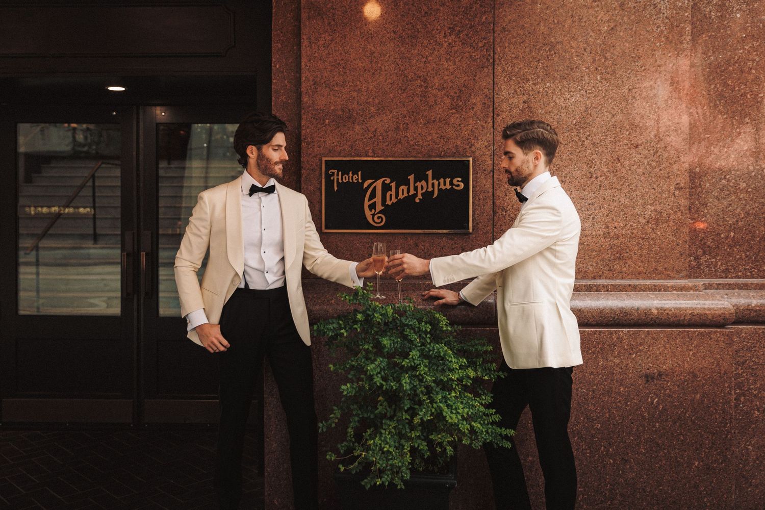 Two grooms in matching white tuxedos toast outside the Hotel Adolphus on their wedding day.