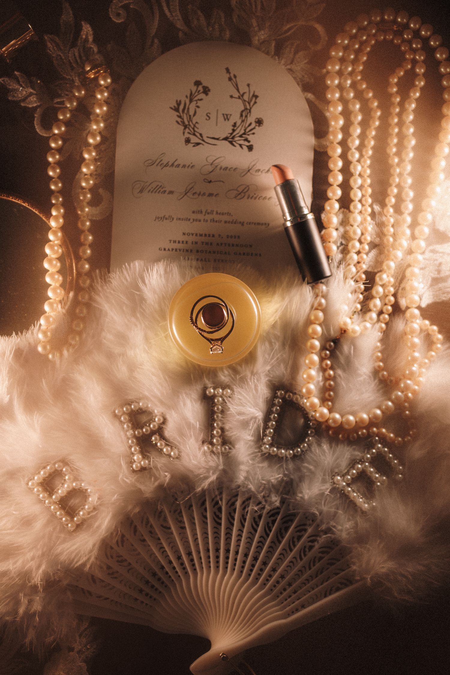 Luxury bridal accessories flat lay featuring pearls, lipstick, wedding invitation, and feather BRIDE fan.