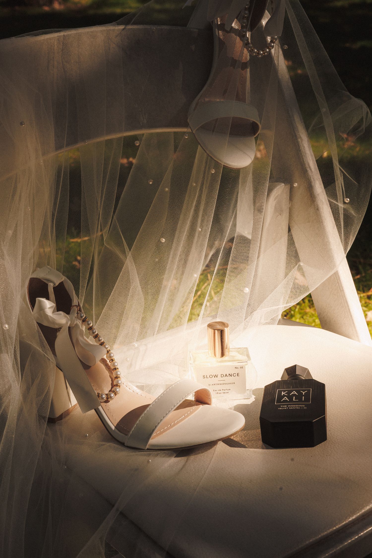 Bridal flat lay with white heels, Slow Dance perfume, Kay Ali fragrance, and a sheer veil draped elegantly.