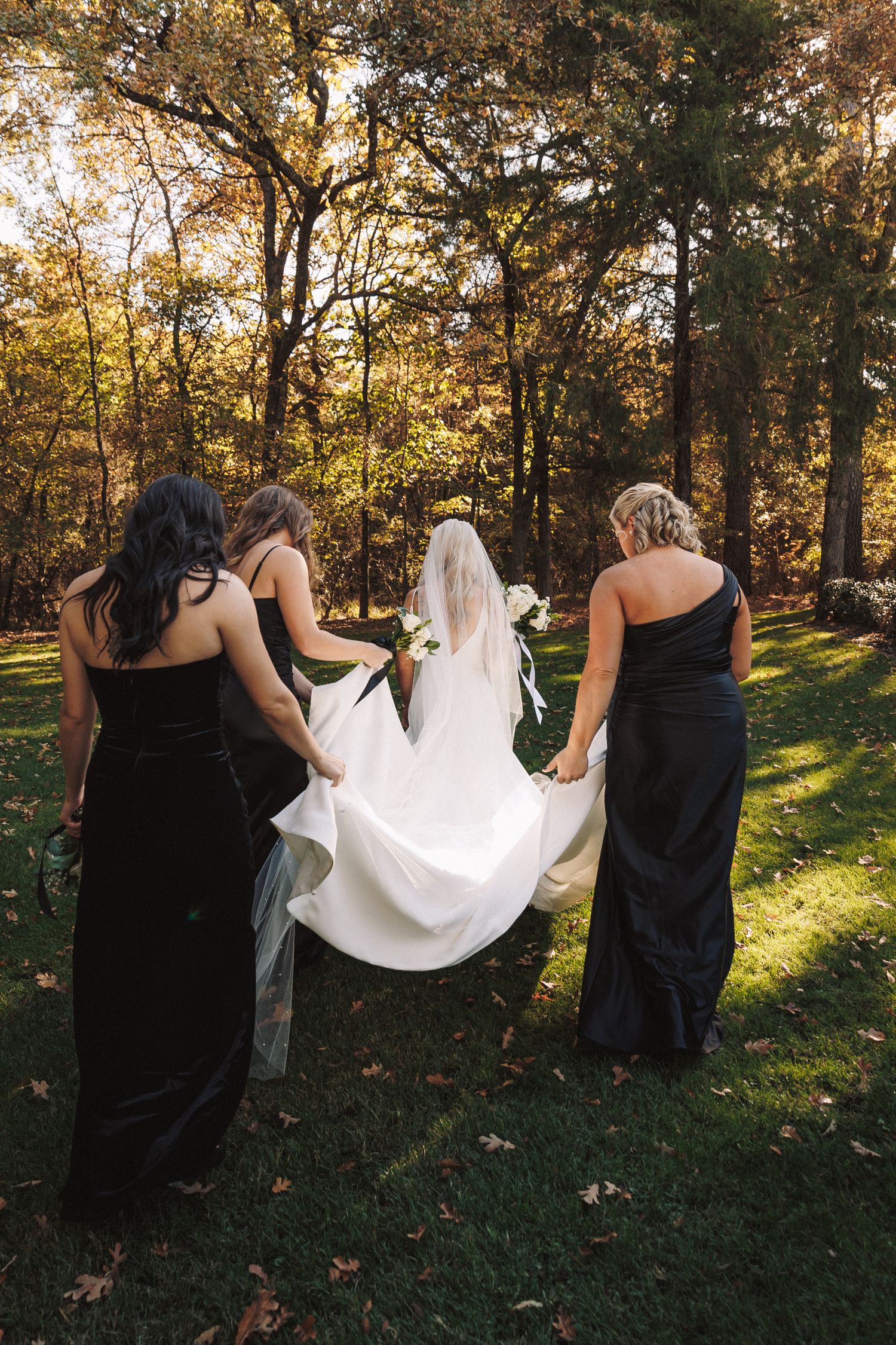 Bridesmaids in black gowns help carry the bride's white wedding dress train as they walk through autumn woodland.
