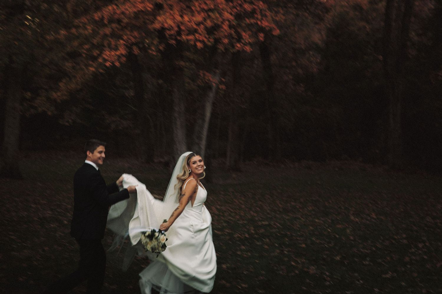 Joyful bride in white gown and veil runs with groom through dramatic autumn red foliage forest.