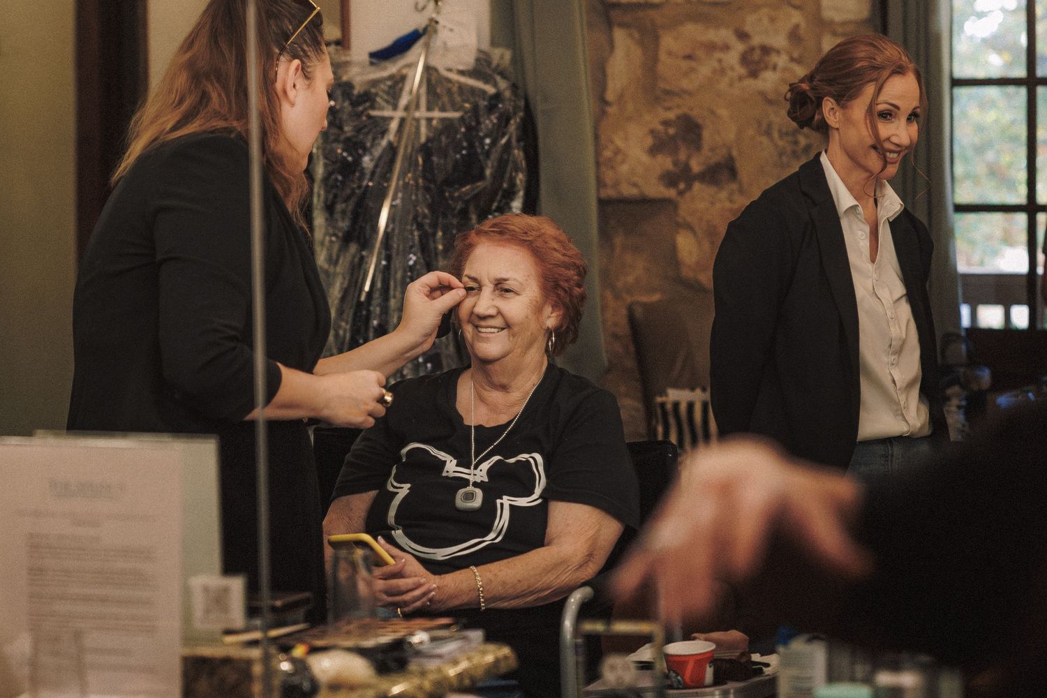 Makeup artist applying eye makeup to a smiling woman at a vanity mirror during wedding prep.