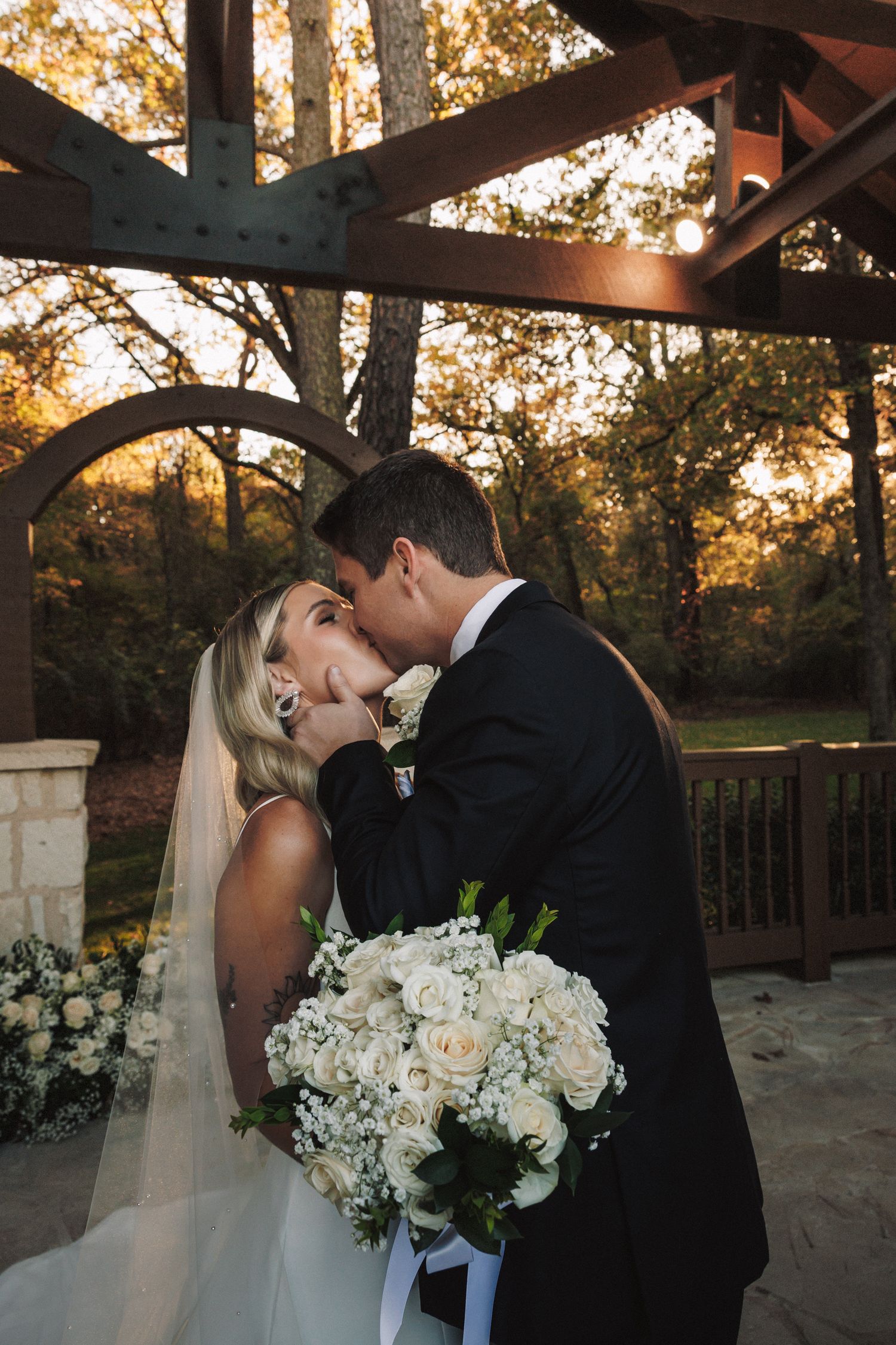 Bride and groom share romantic kiss under rustic wooden pergola at golden hour with white roses.