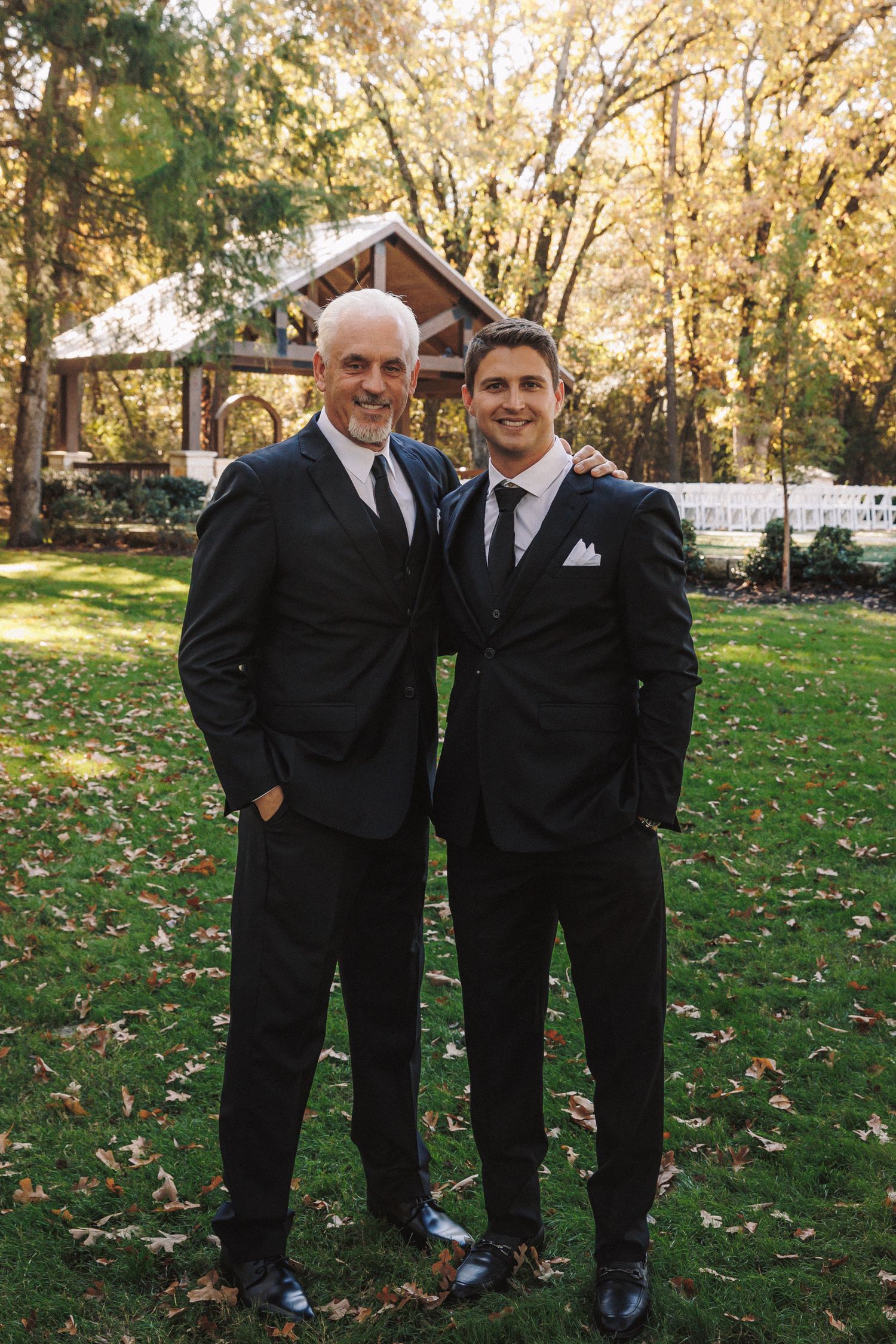 Two men in black suits and ties pose together outdoors at a fall wedding venue.