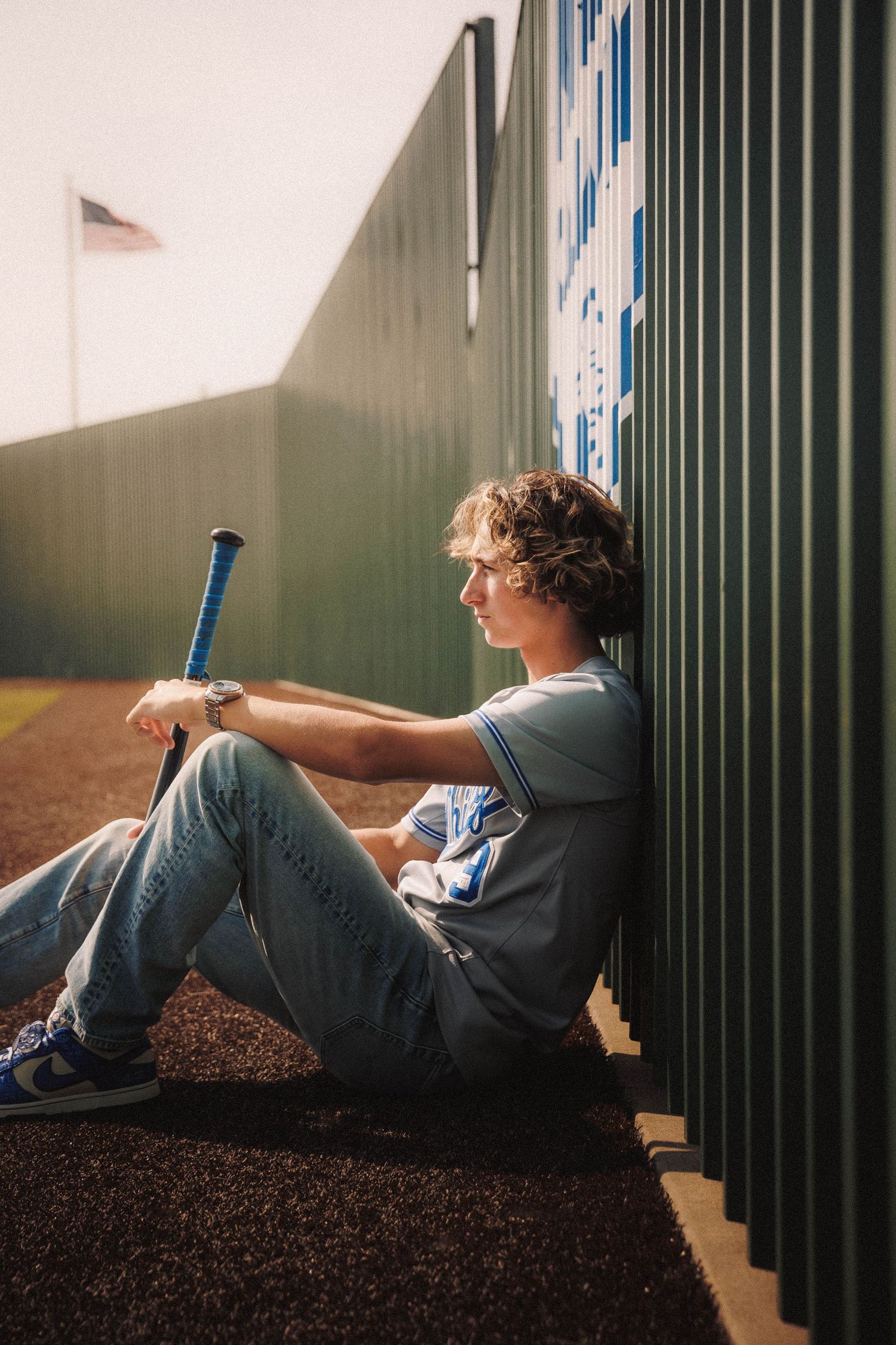 Baseball player in gray t-shirt and jeans sits against fence at field during sunset holding bat.