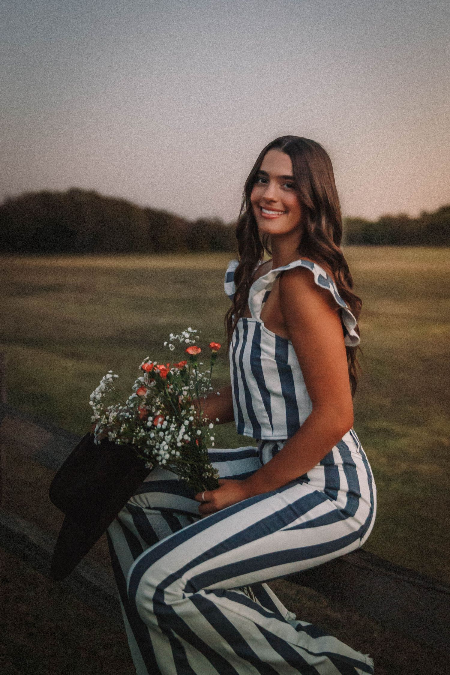 Woman in striped jumpsuit poses outdoors at sunset with flowers and golden grass in the background.