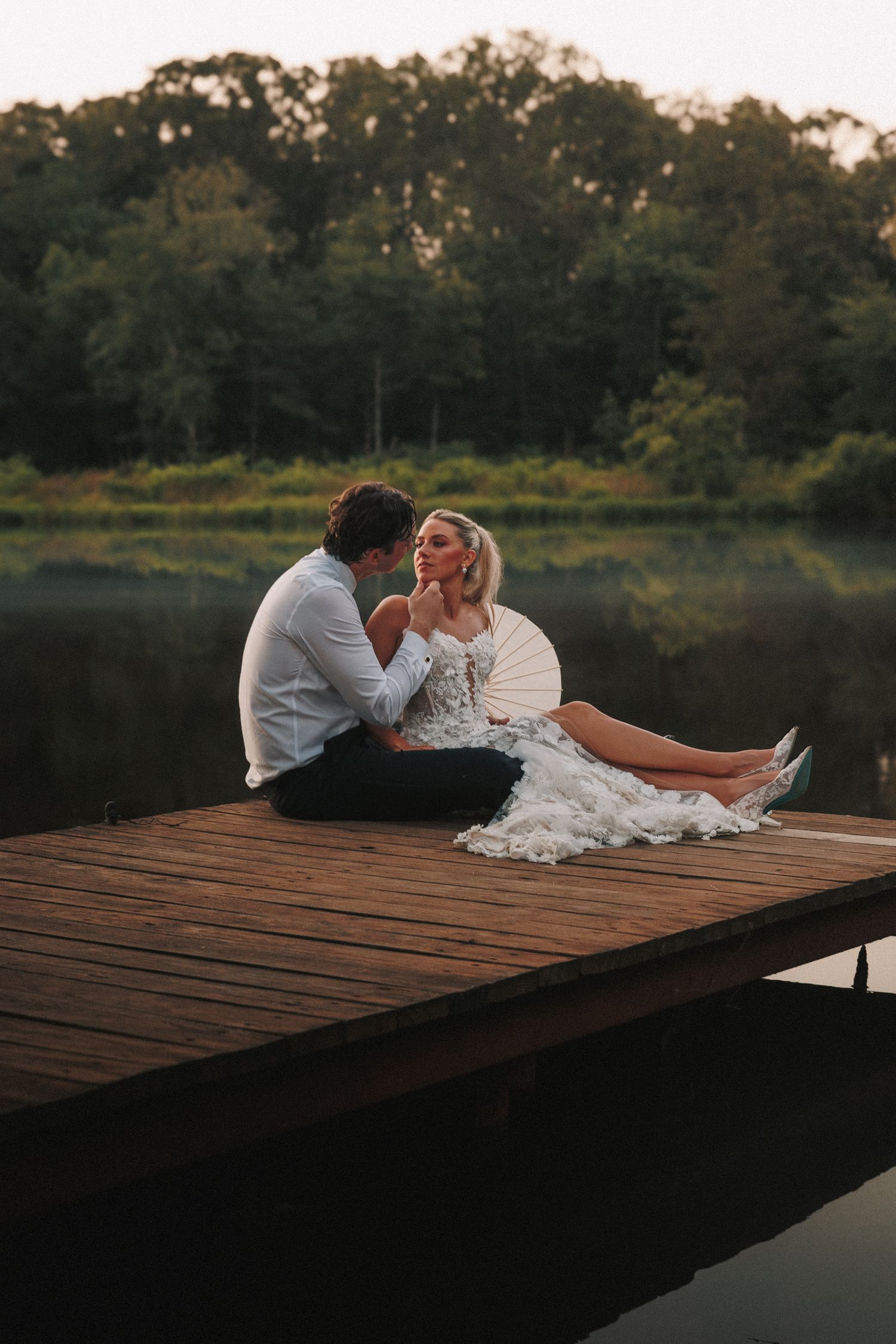 A couple sits intimately on a wooden dock by a serene lake at dusk surrounded by trees.