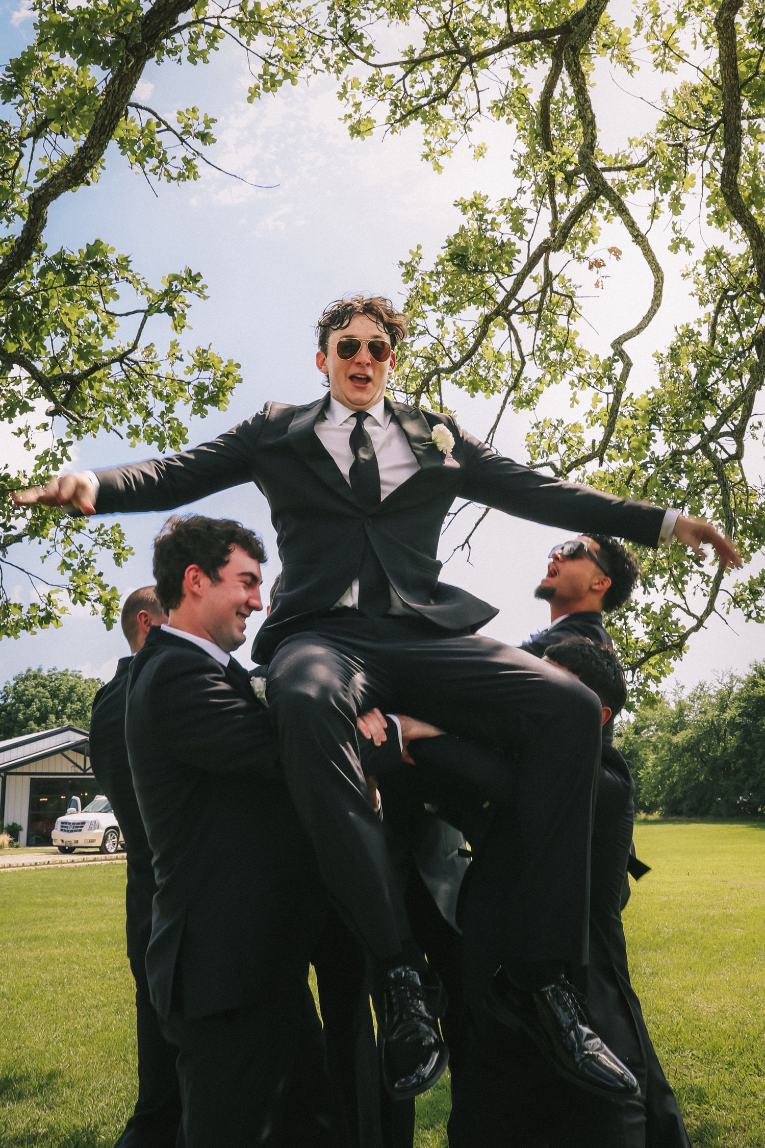 Groomsmen in black suits celebrate outdoors under tree branches on a sunny day.