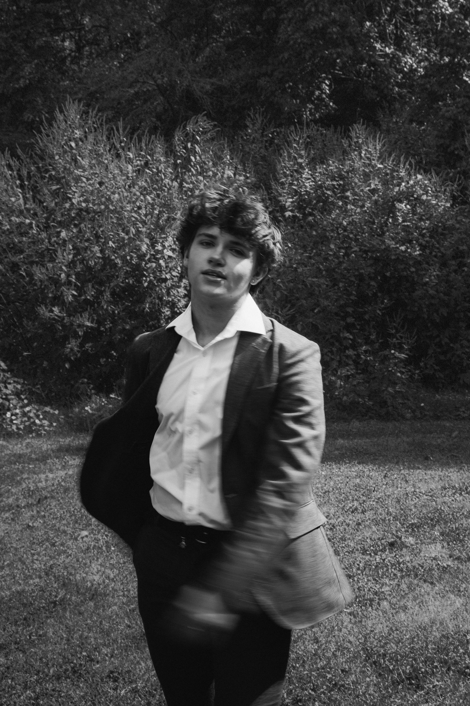 Black and white portrait of someone wearing a dark suit and white shirt posing outdoors in a garden setting.